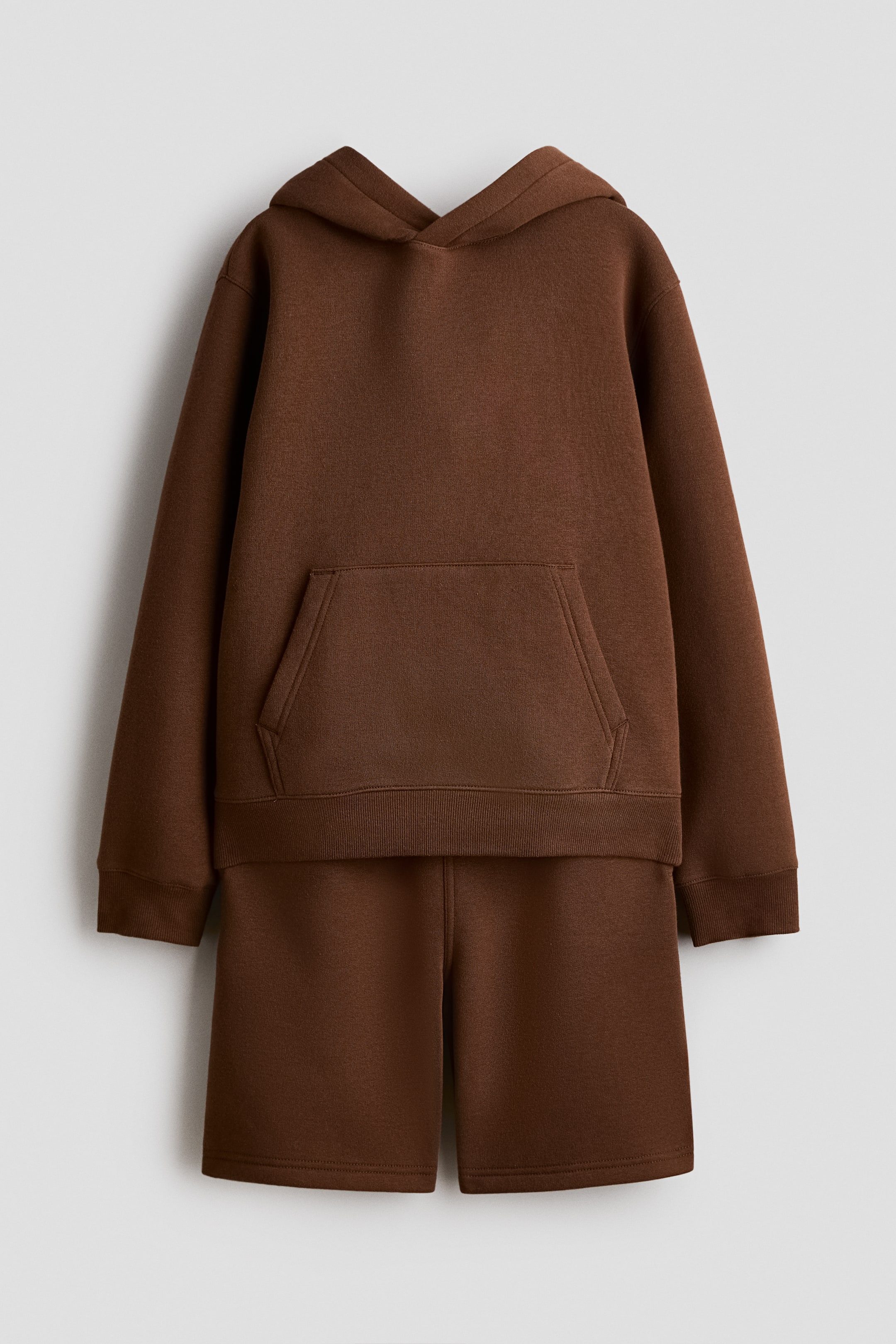 Näytä suurempi kuva: Ruskea sweatshirt-setti on esitetty FRONT-näkymässä; siihen kuuluu hoodie ja sopivat shortsit. Hoodiessa on vuorattu, wrapover-front-huppu, kangaroo pocket sekä resorit hihansuissa ja helmassa, kun taas shortseissa on sileä vyötärönauha ja ne yltävät reiden puoliväliin.