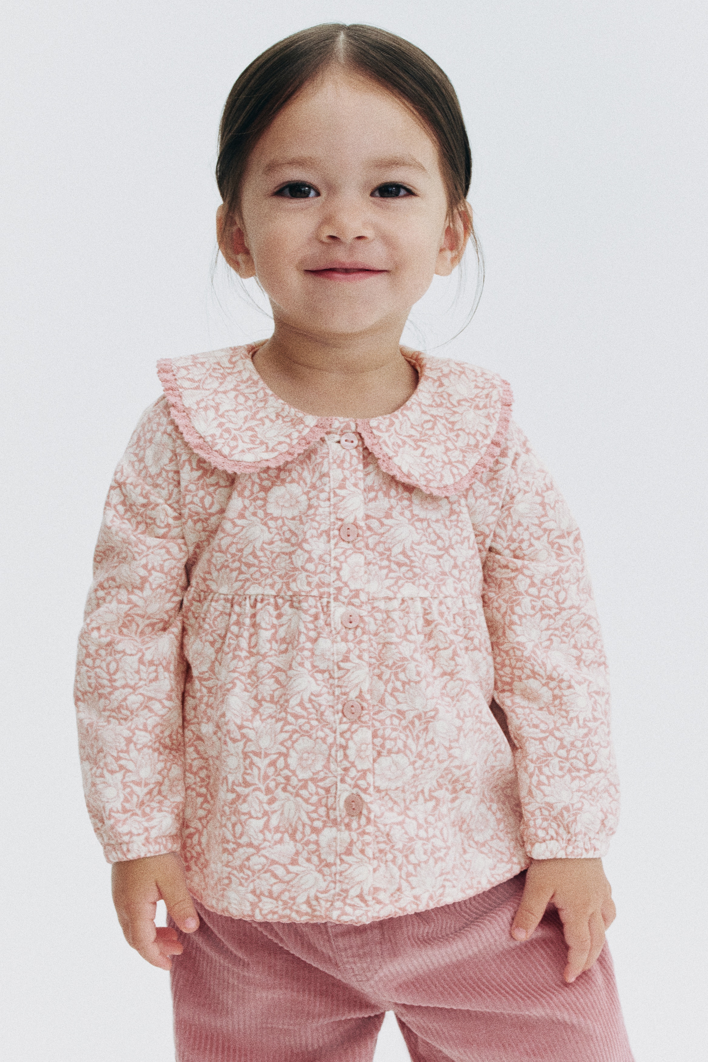Cette vue de face met en valeur une blouse en velours côtelé à motif floral rose et blanc, dotée dâun col Claudine bordé de dentelle et de boutons assortis sur le devant. Le design présente une couture froncée au-dessus de la taille pour plus de volume, ainsi que des manches longues avec poignets élastiqués.