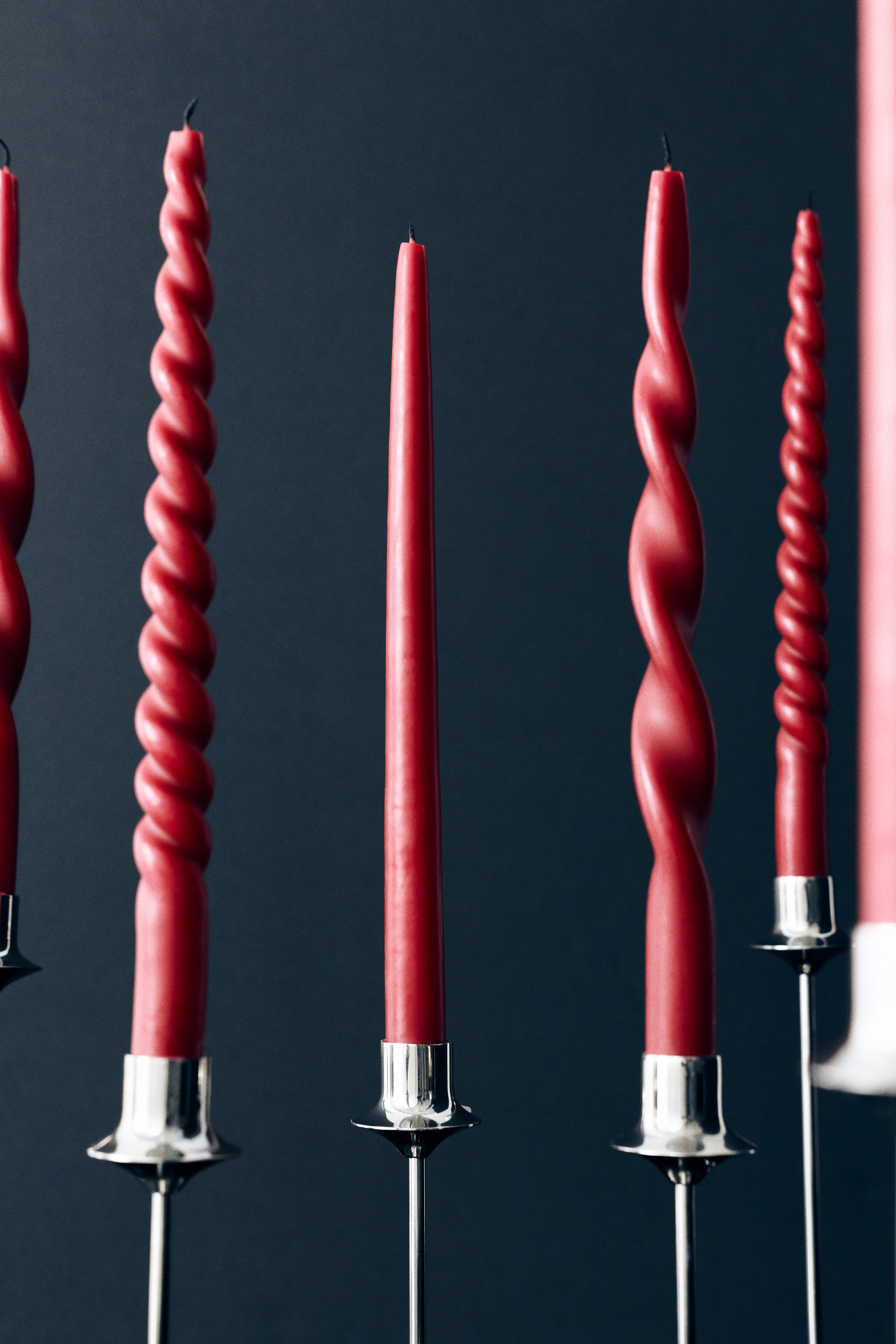 Three pairs of red taper candles are hanging side-by-side from continuous wicks, displaying three distinct shapes from left to right: a tight spiral, a loose spiral, and a classic smooth taper.