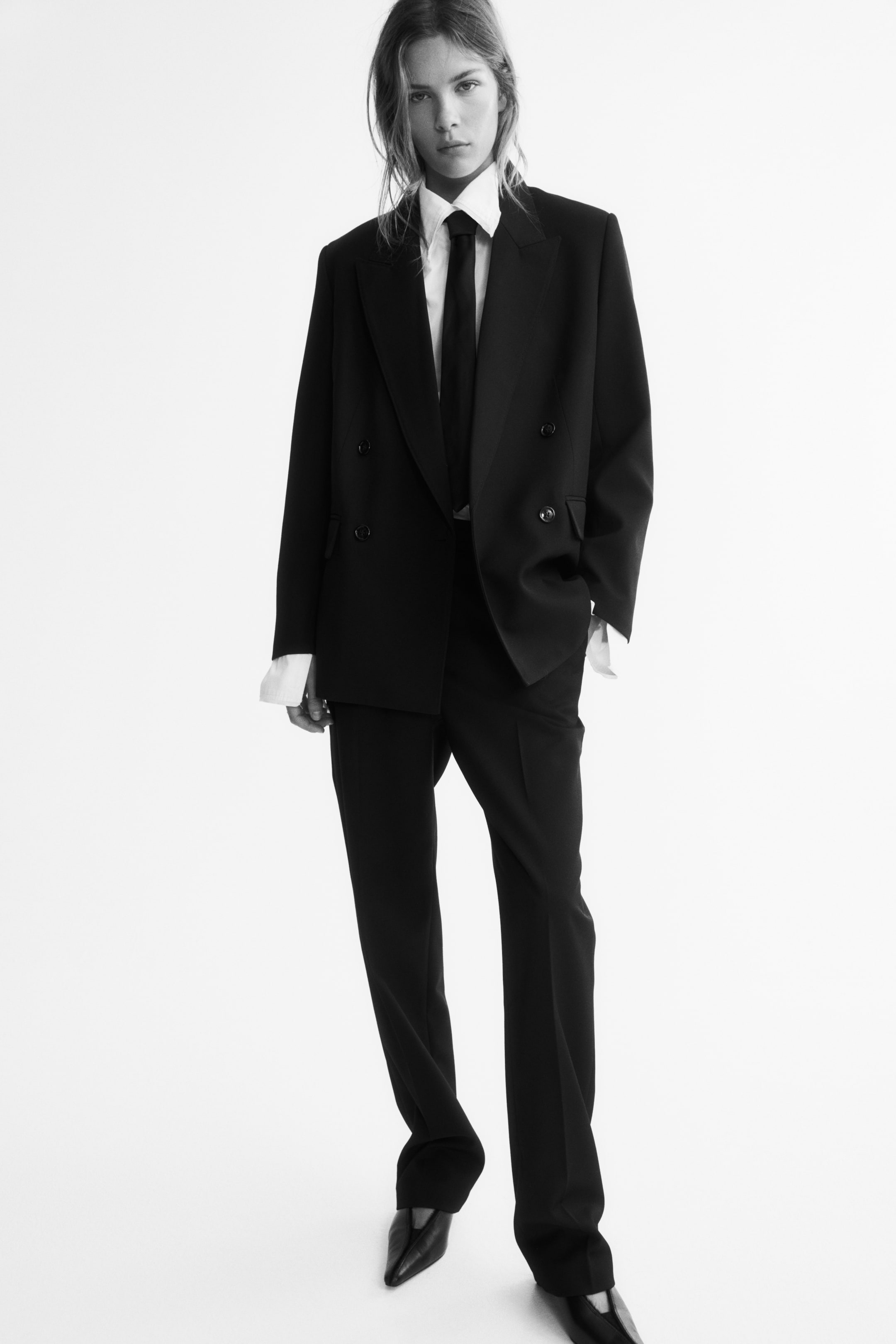 View larger image: Dark trousers featuring slim legs and pressed creases down the front are being worn by the model. The trousers are styled with a matching double-breasted blazer, a white collared shirt, a dark tie, and pointed-toe shoes.