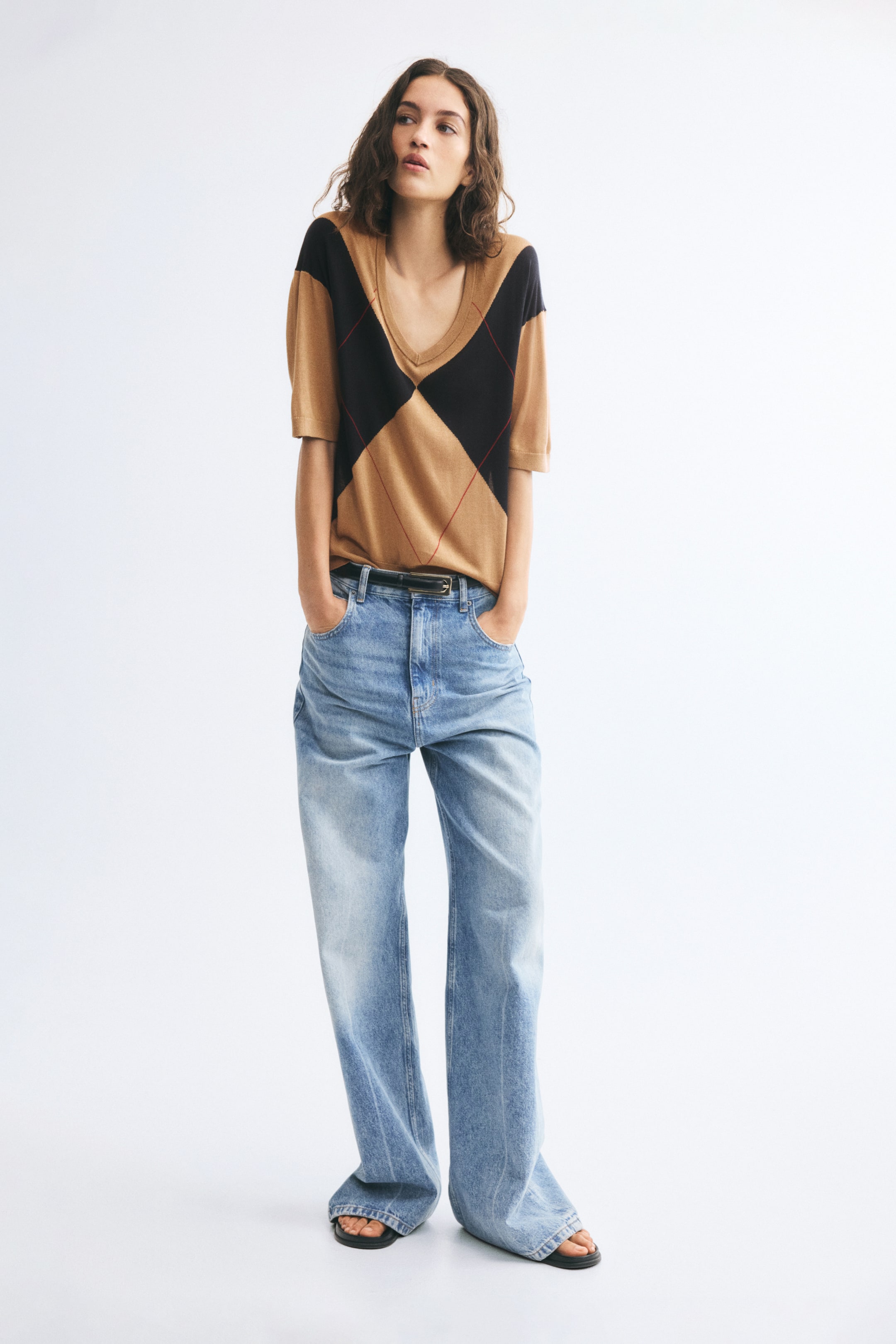 View larger image: An argyle-patterned intarsia knit top in brown, black, and red is worn by the model, tucked into light-blue jeans. The top features a deep, ribbed V-neck, dropped shoulders, and short sleeves that end just above the elbows.