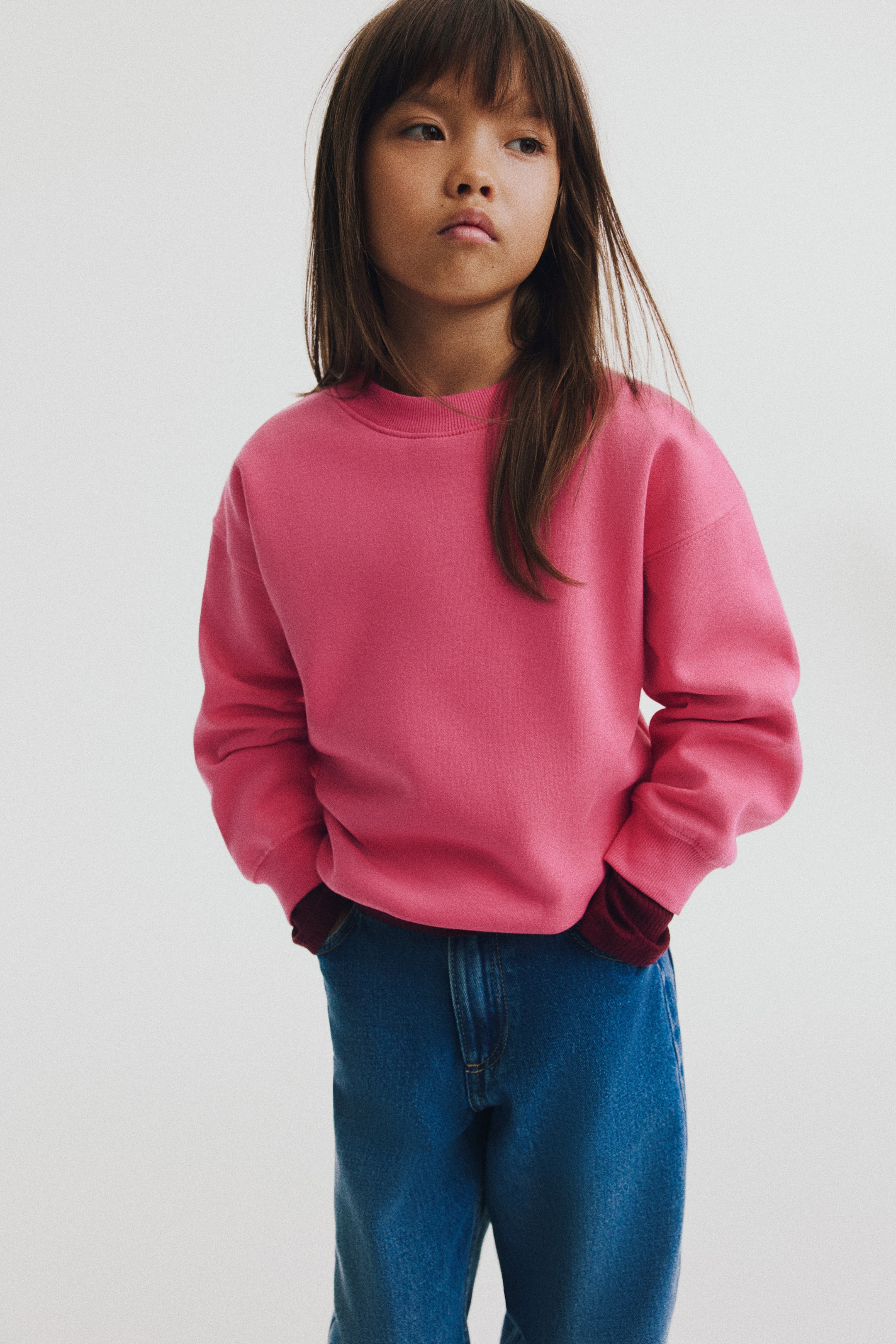 View larger image: A pink lightweight sweatshirt is shown from the front, featuring a round ribbed neckline, dropped shoulders, and long sleeves with ribbed cuffs. The relaxed-fit sweater is styled layered over a dark burgundy long-sleeve top and paired with blue jeans.