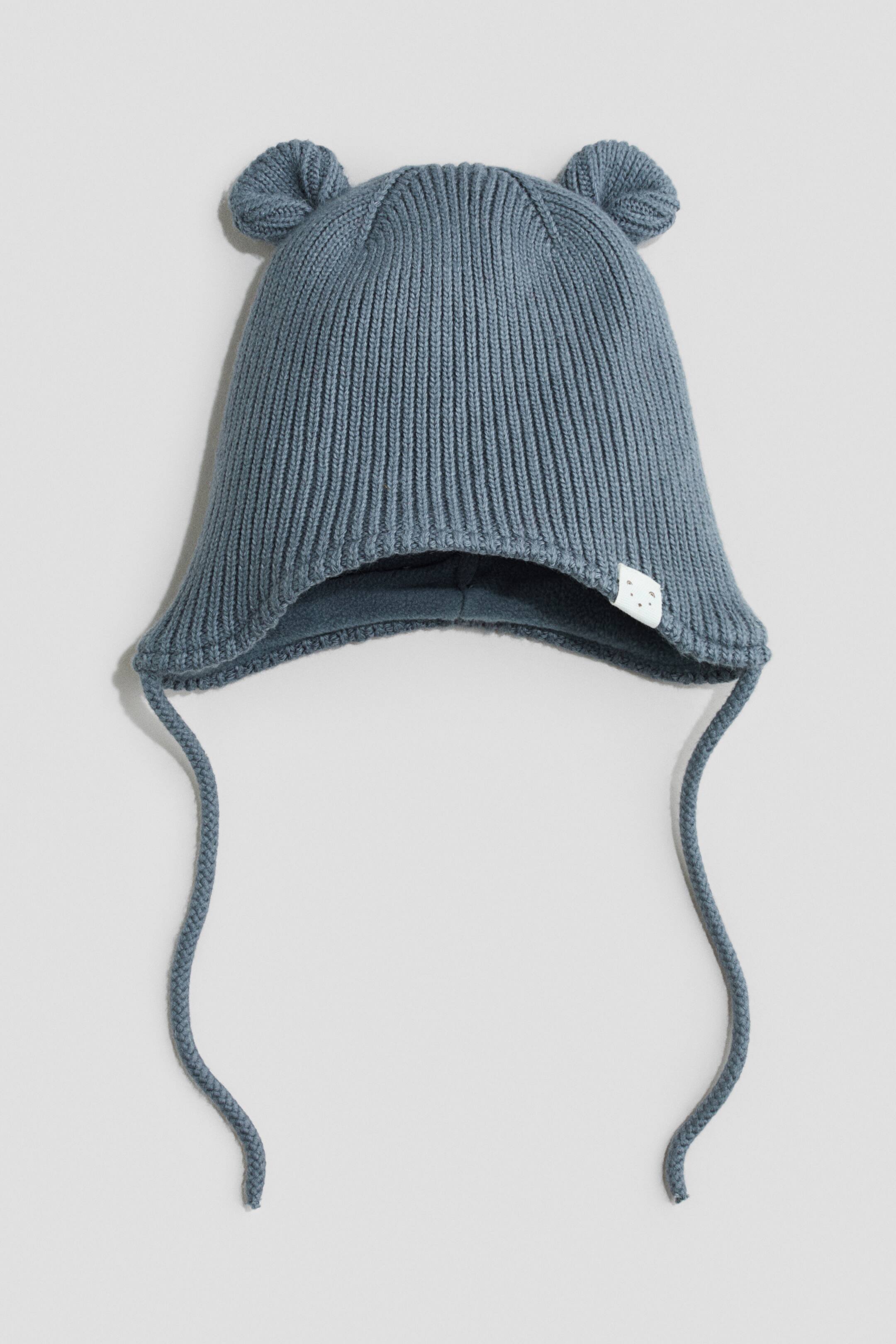 View larger image: A blue-grey fleece-lined earflap beanie is shown from the front, highlighting a ribbed-knit texture and two small decorative ears on the crown. Long, thin knit ties extend from the earflaps, while a matching fleece lining is visible on the inside.