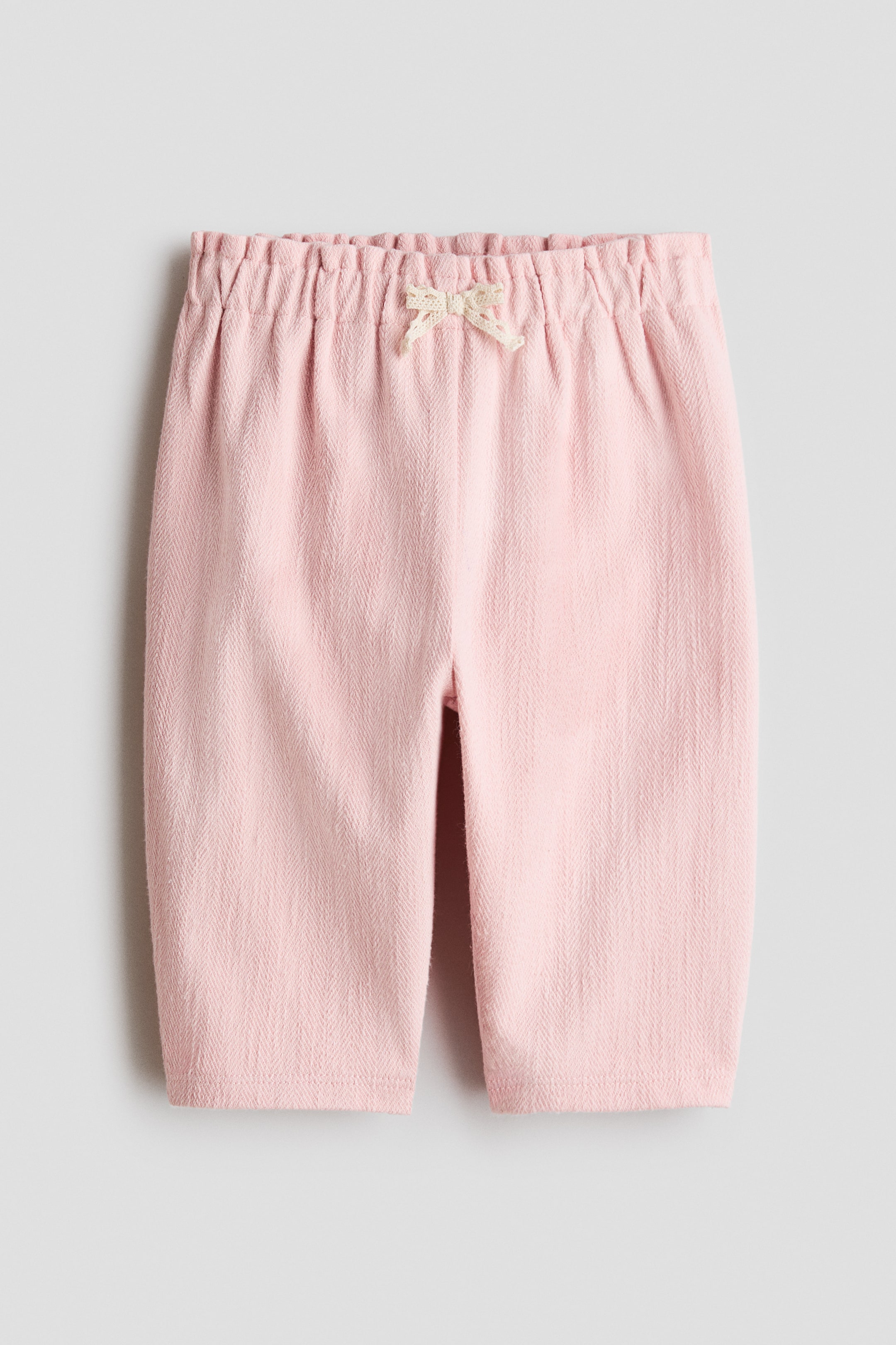 Ampliar la imagen: El frente de los pantalones rosas presenta una pretina elástica fruncida con un pequeño lazo de color claro. Los pantalones muestran una sutil textura de espiga.