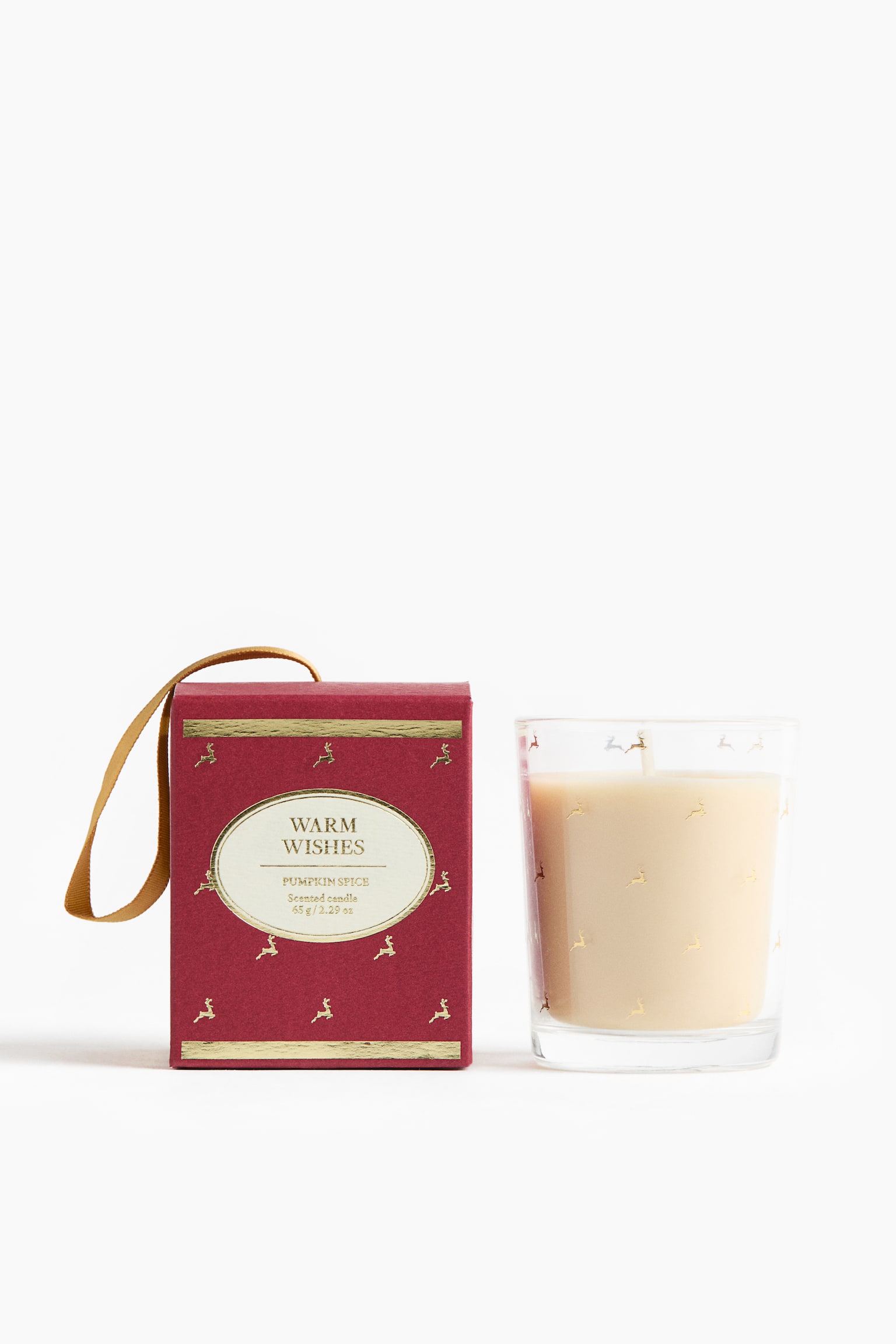 Una vela aromÃ¡tica y su caja de regalo estÃ¡n colocadas una junto a la otra, de frente. La caja rectangular roja de la izquierda presenta un lazo dorado, un patrÃ³n de pequeÃ±os renos dorados y una etiqueta ovalada color crema que dice "WARM WISHES PUMPKIN SPICE". A la derecha, la vela de vidrio transparente contiene cera color crema y muestra motivos de renos dorados a juego.