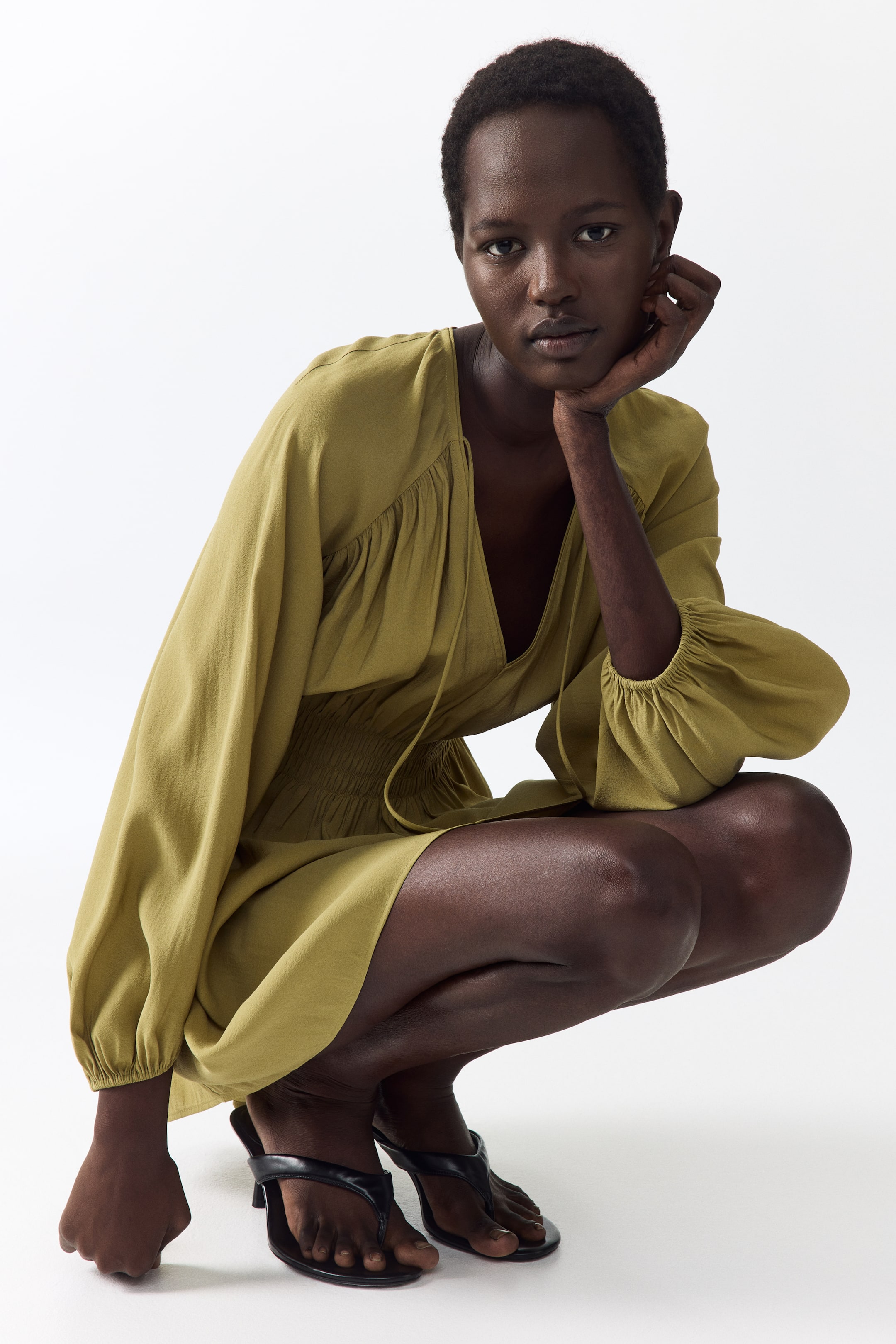 View larger image: An olive green mini dress featuring a deep V-neckline with a drawstring tie, gathered detailing at the shoulders, long voluminous sleeves with elasticated cuffs, and a shirred elasticated waist.