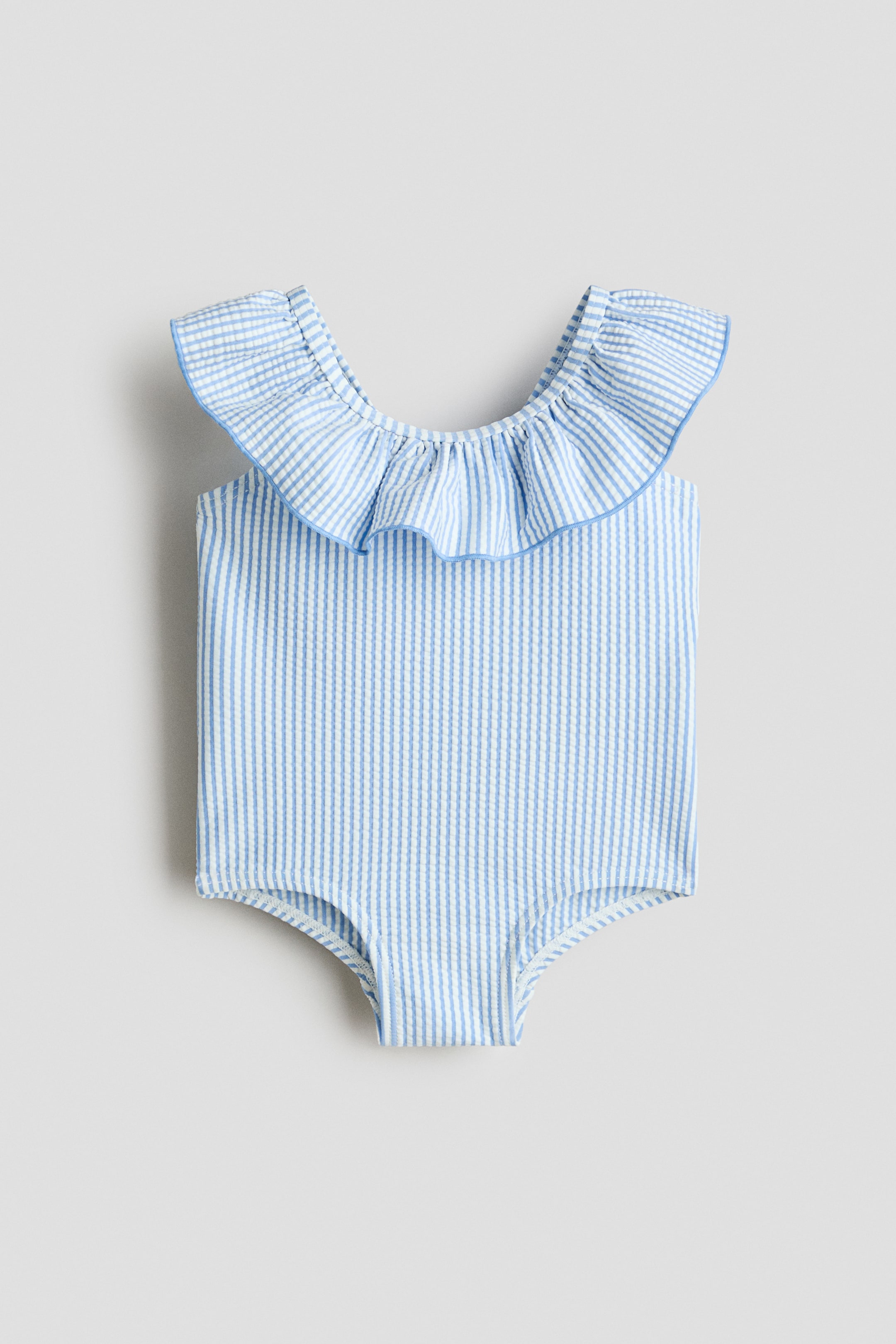 View larger image: The baby's one-piece swimsuit features a light blue and white seersucker vertical stripe pattern. A wide, gathered ruffle forms the neckline and extends over the shoulder areas, edged with a solid light blue piping.