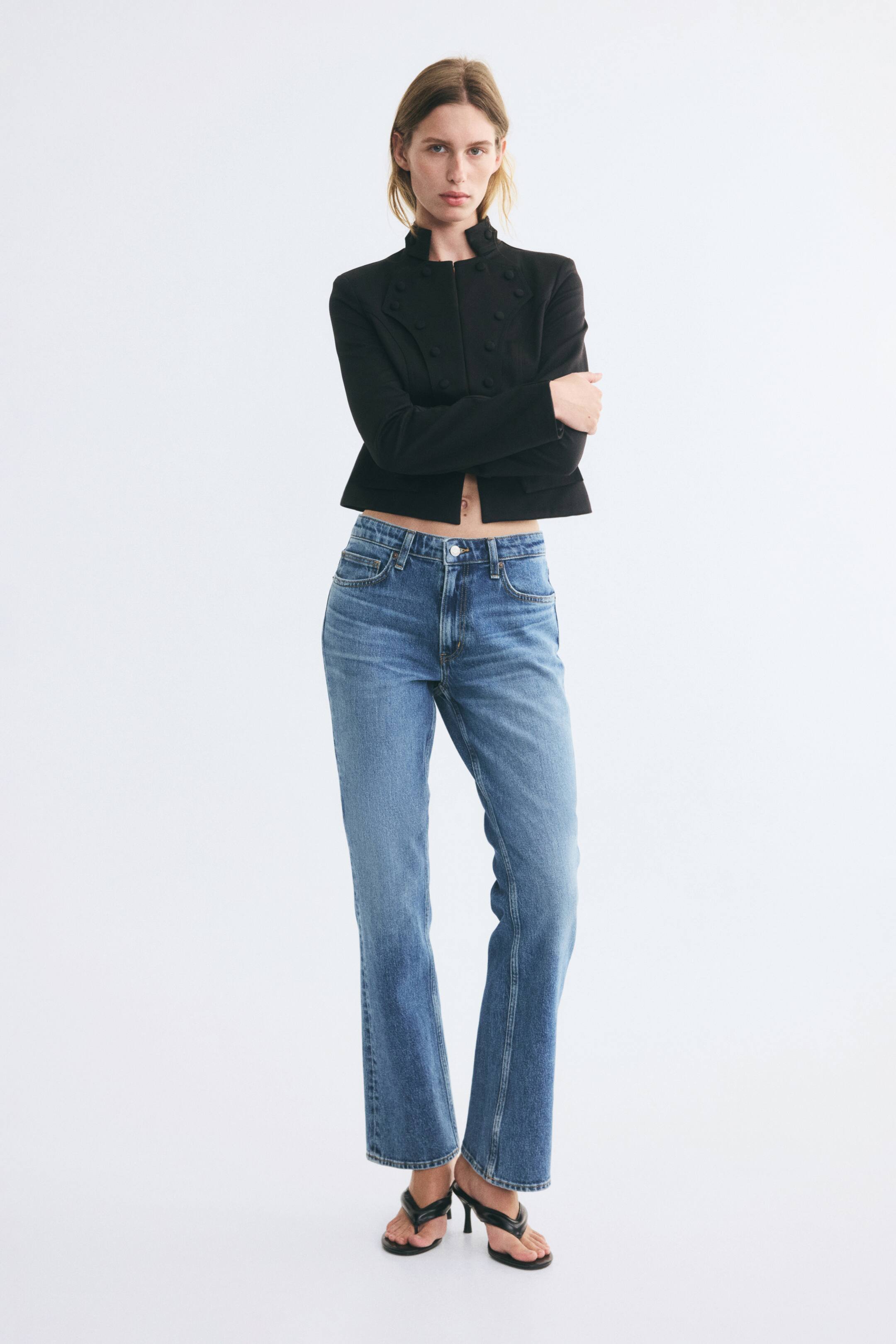 Ver imagen más grande: La modelo lleva unos vaqueros de denim azul de cinco bolsillos, con cintura regular, corte slim en los muslos y un ligero bajo acampanado tipo bootcut. Los vaqueros se combinan con una chaqueta negra corta y sandalias negras de tacón.