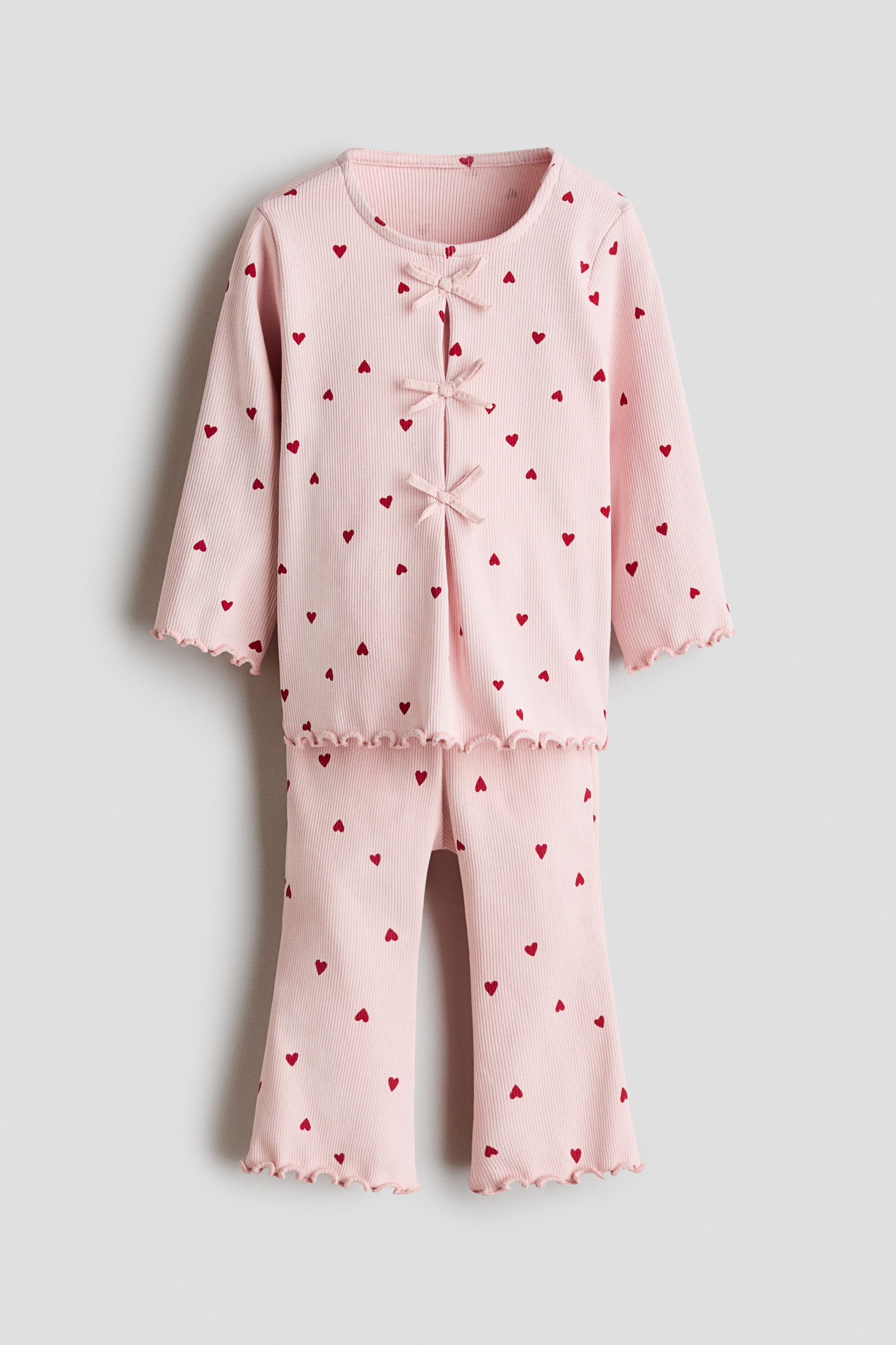 View larger image: A pink pajama set is shown from the front, featuring a ribbed knit top and matching pants, both covered in a pattern of small red hearts. The top has a round neckline, long sleeves with ruffled cuffs, a ruffled hem, and three decorative bows down the front, while the pants have long legs with ruffled hems.