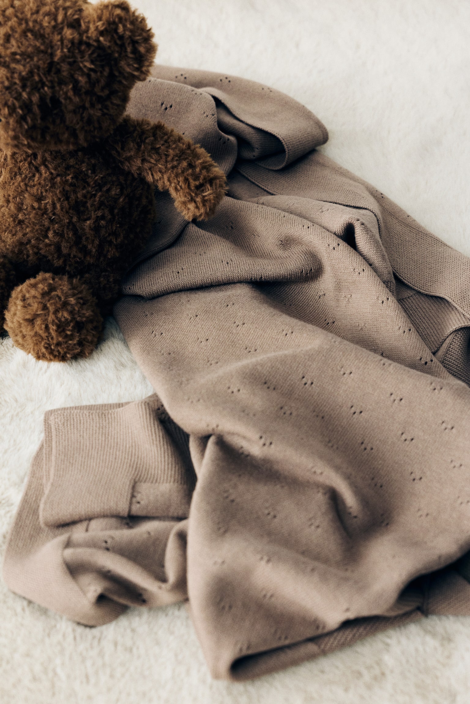 Дитяча ковдра з м’якого бавовняного pointelle knit у приглушеному тауповому відтінку лежить, вільно драпірована, з делікатним повторюваним візерунком eyelet і широкою ребристою облямівкою. Коричневий плюшевий ведмедик teddy bear спочиває у верхньому лівому куті складеної ковдри. - 1