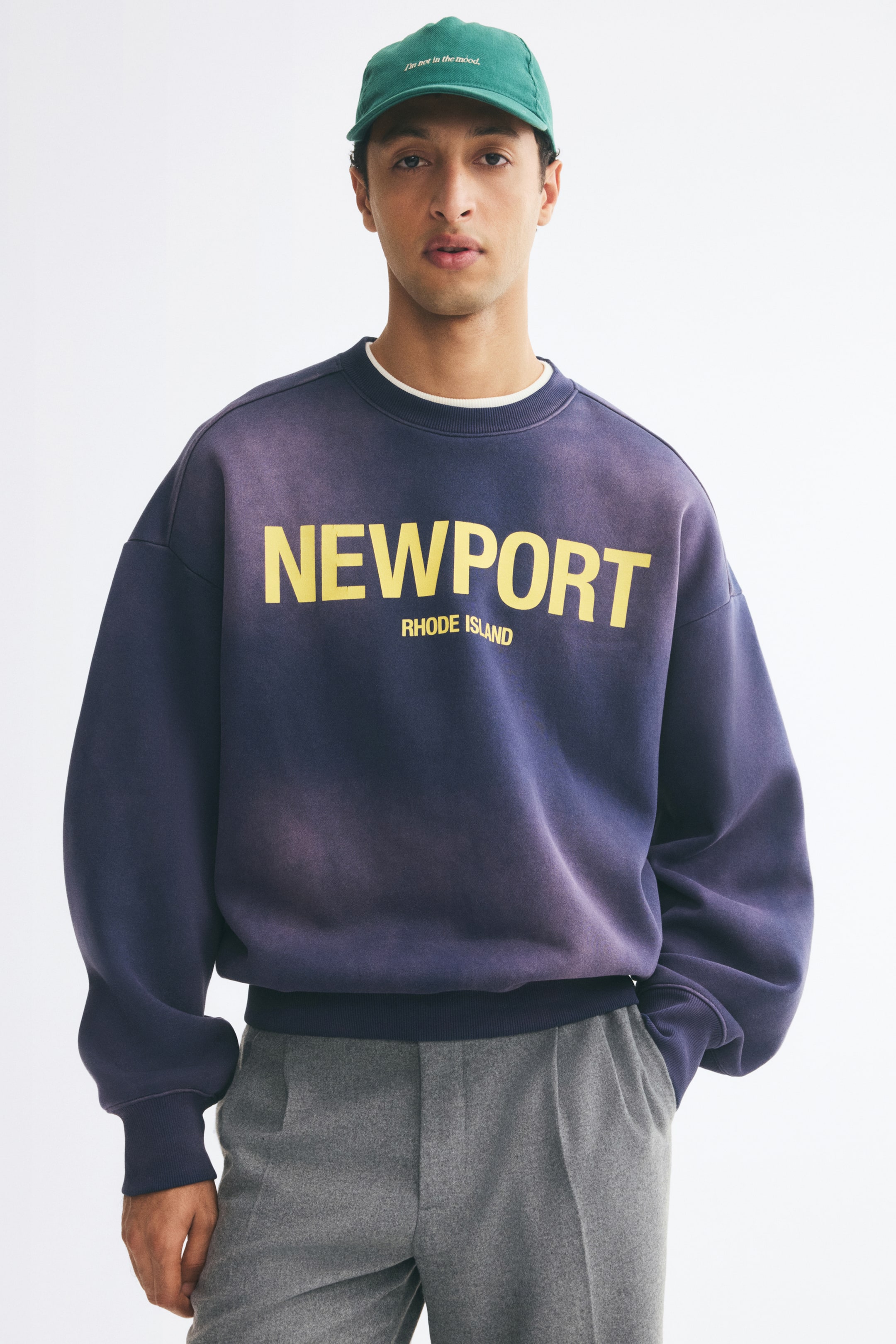 View larger image: An oversized, faded purple-blue sweatshirt featuring a yellow "NEWPORT RHODE ISLAND" graphic is being worn by the model. The sweatshirt has dropped shoulders and ribbed trims, and is layered over a white T-shirt and styled with grey trousers.