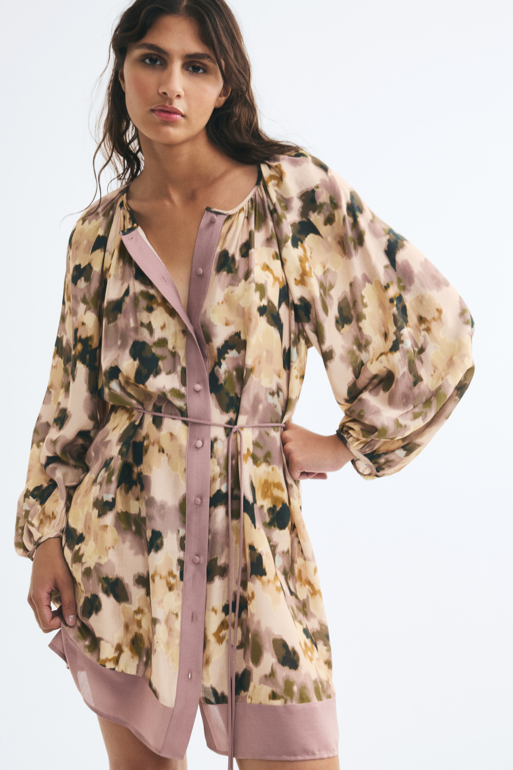 View larger image: The model is wearing a short, oversized tunic dress with an abstract pattern in beige, olive green, and mauve. The dress features a solid mauve button-front placket, long balloon sleeves, a matching slim tie belt at the waist, and a solid trim at the hem.