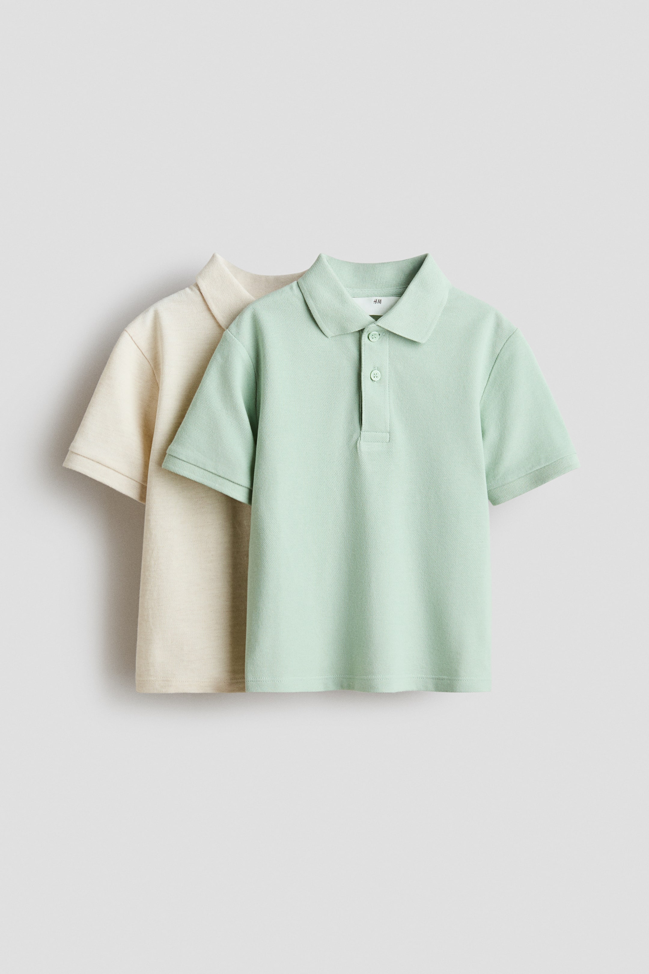 이미지 확대 보기: 두 벌의 폴로 셔츠가 정면을 향해 진열되어 있으며, 밝은 베이지색 셔츠 앞에 밝은 연두색 셔츠가 레이어드되어 있습니다. 앞쪽의 밝은 연두색 폴로 셔츠는 클래식 칼라, 2버튼 플래킷, 소맷단이 시보리 처리된 반소매를 특징으로 합니다.