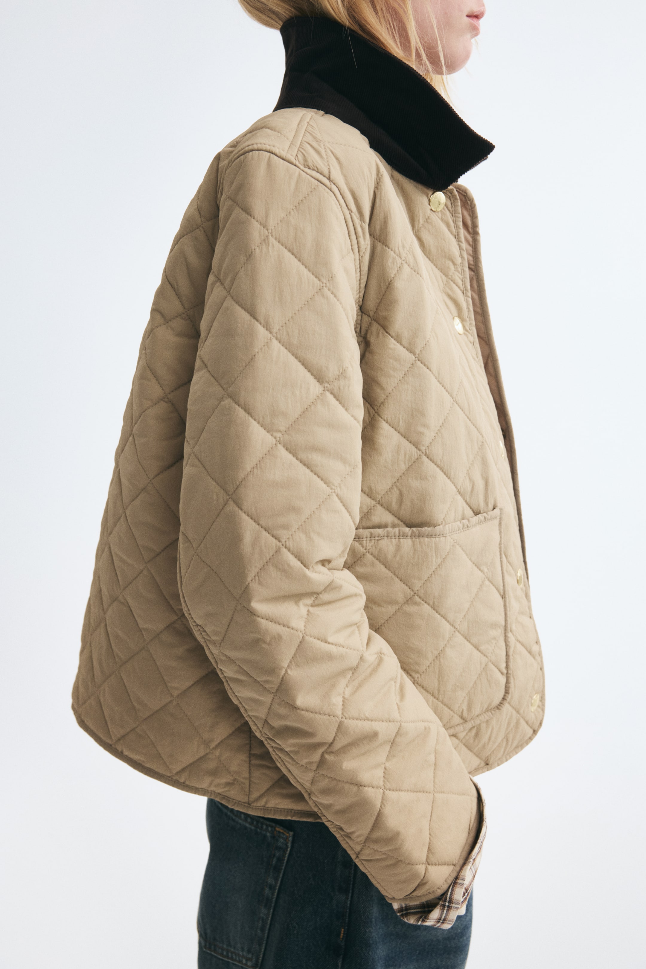 Visualizza l'immagine ingrandita: Una giacca trapuntata beige con motivo a rombi viene indossata dalla modella, mostrata di profilo. La giacca presenta un colletto in velluto a coste marrone scuro, bottoni a pressione dorati lungo il davanti, una tasca applicata e una fodera a quadri visibile al polsino, abbinata a jeans blu scuro.