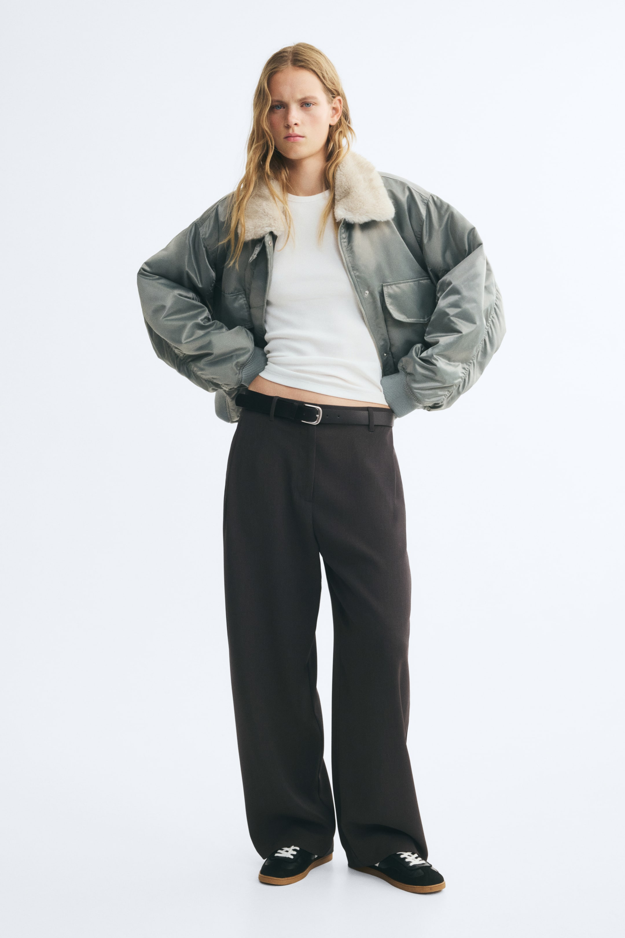 Visualizza l'immagine ingrandita: Pantaloni a vestibilità morbida in tessuto intrecciato marrone scuro indossati dalla modella, con vita regolare, pieghe frontali e cintura nera con fibbia in metallo. I pantaloni sono abbinati a un top bianco, una giacca bomber grigia con colletto in pelliccia sintetica e sneakers nere.