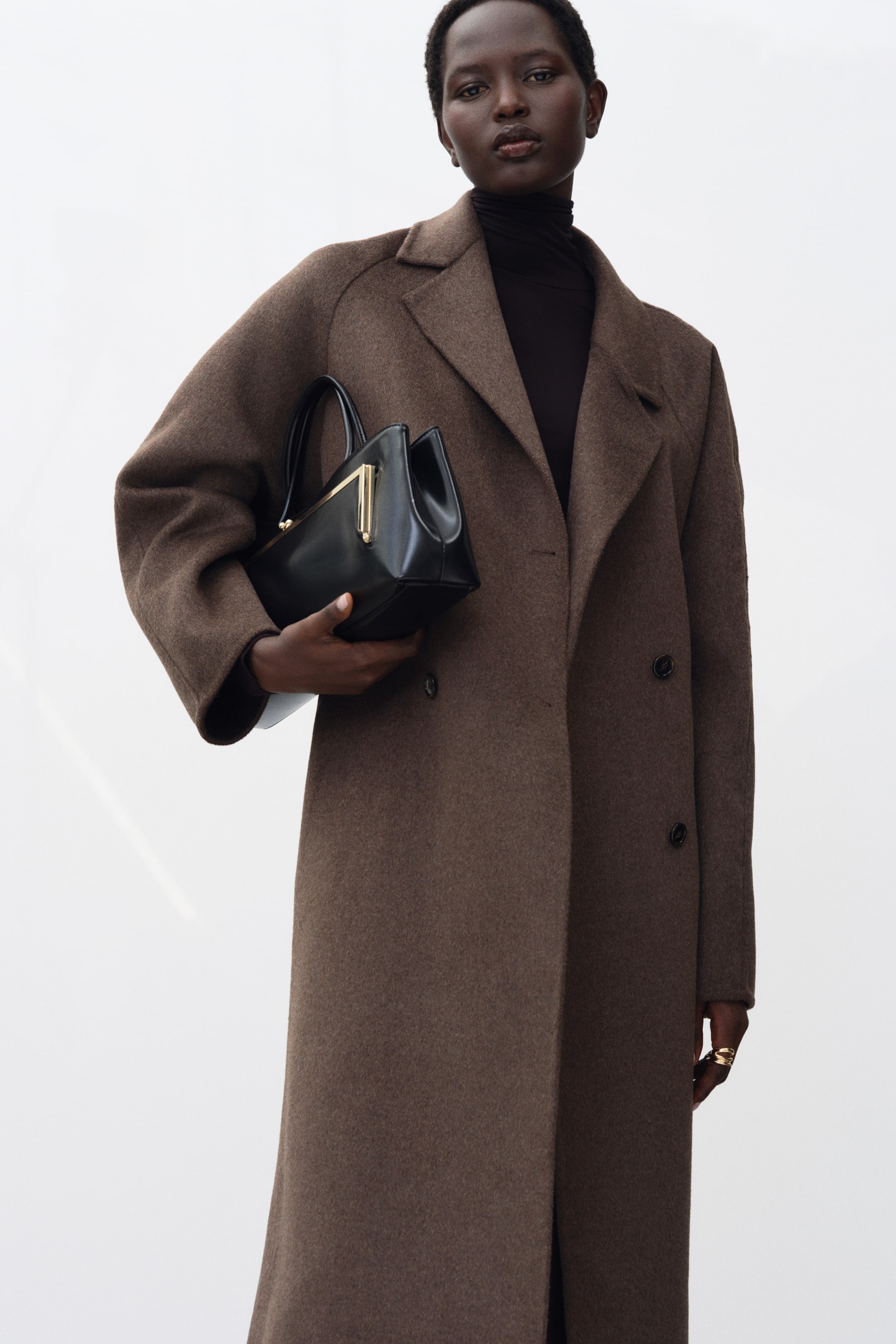 View larger image: A long brown wool-blend coat featuring notch lapels, raglan sleeves, and double-breasted button fasteners is worn by the model over a black turtleneck. The model is holding a structured black handbag with gold-toned hardware under one arm.