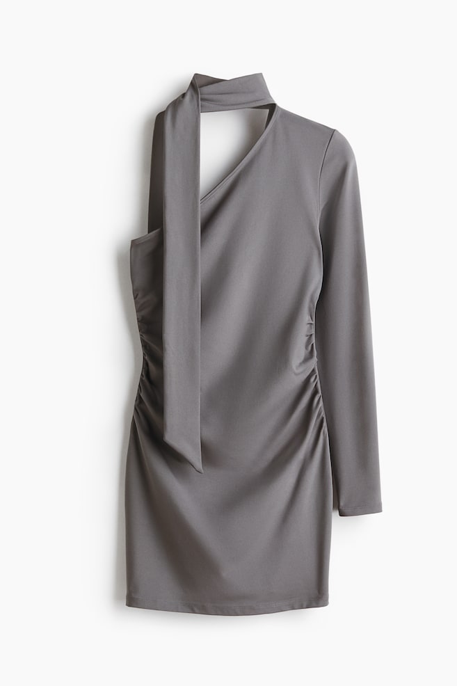 Une robe mini grise est présentée de FACE, avec une encolure asymétrique comportant un col en V à droite et un col haut à gauche, d'où une longue cravate descend en drapé. La robe présente également une manche longue à droite et un froncage extensif le long de son côté gauche. - 1