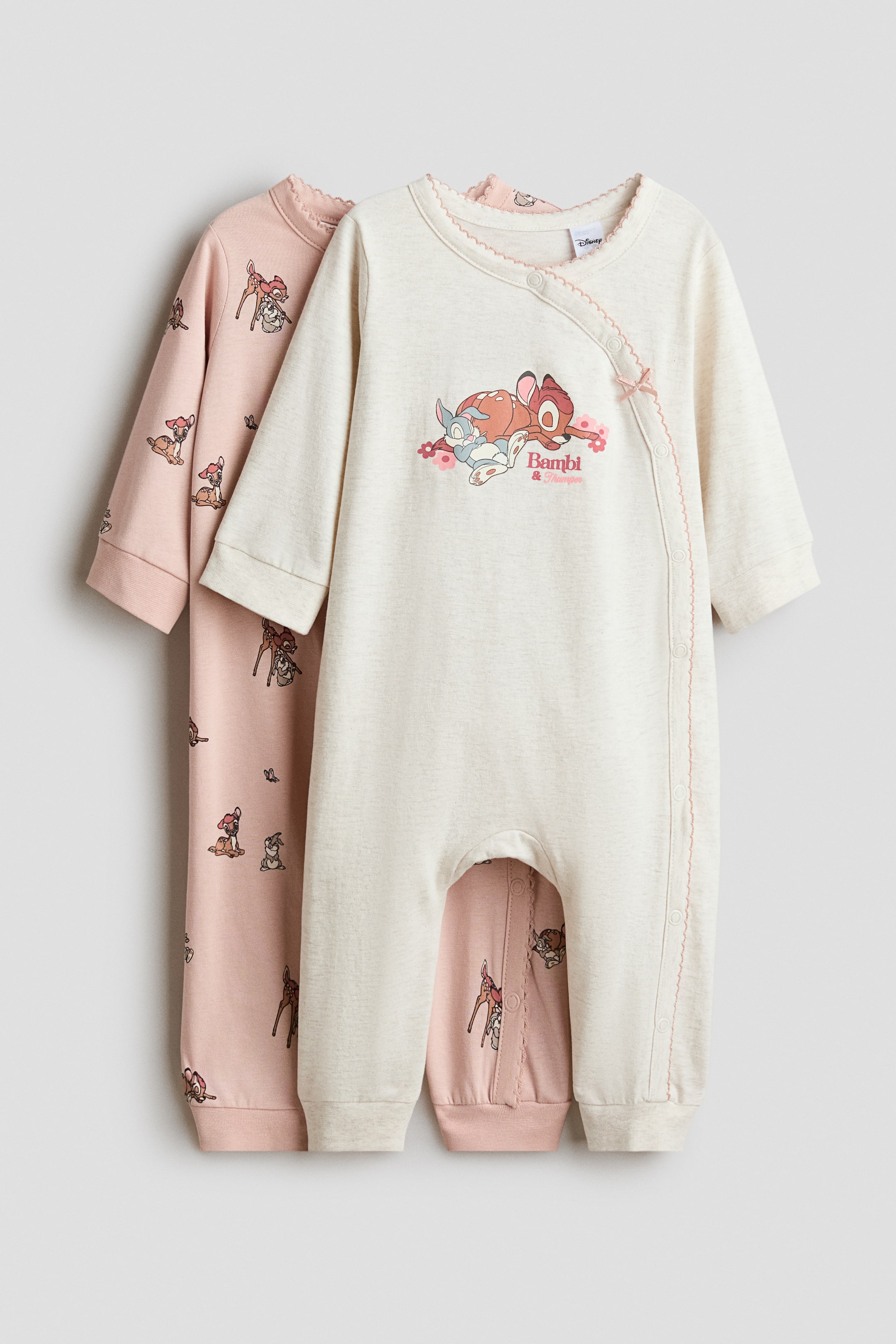 View larger image: Two baby rompers are on display, one in off-white and one in pink, both featuring long sleeves and full-length legs with ribbed cuffs. The off-white romper, positioned in front, features a graphic print of Bambi and Thumper on the chest, a diagonal snap-button closure accented with a small pink bow, and a scalloped neckline. The pink romper, partially visible behind, is patterned with an all-over print of smaller Bambi and Thumper characters.