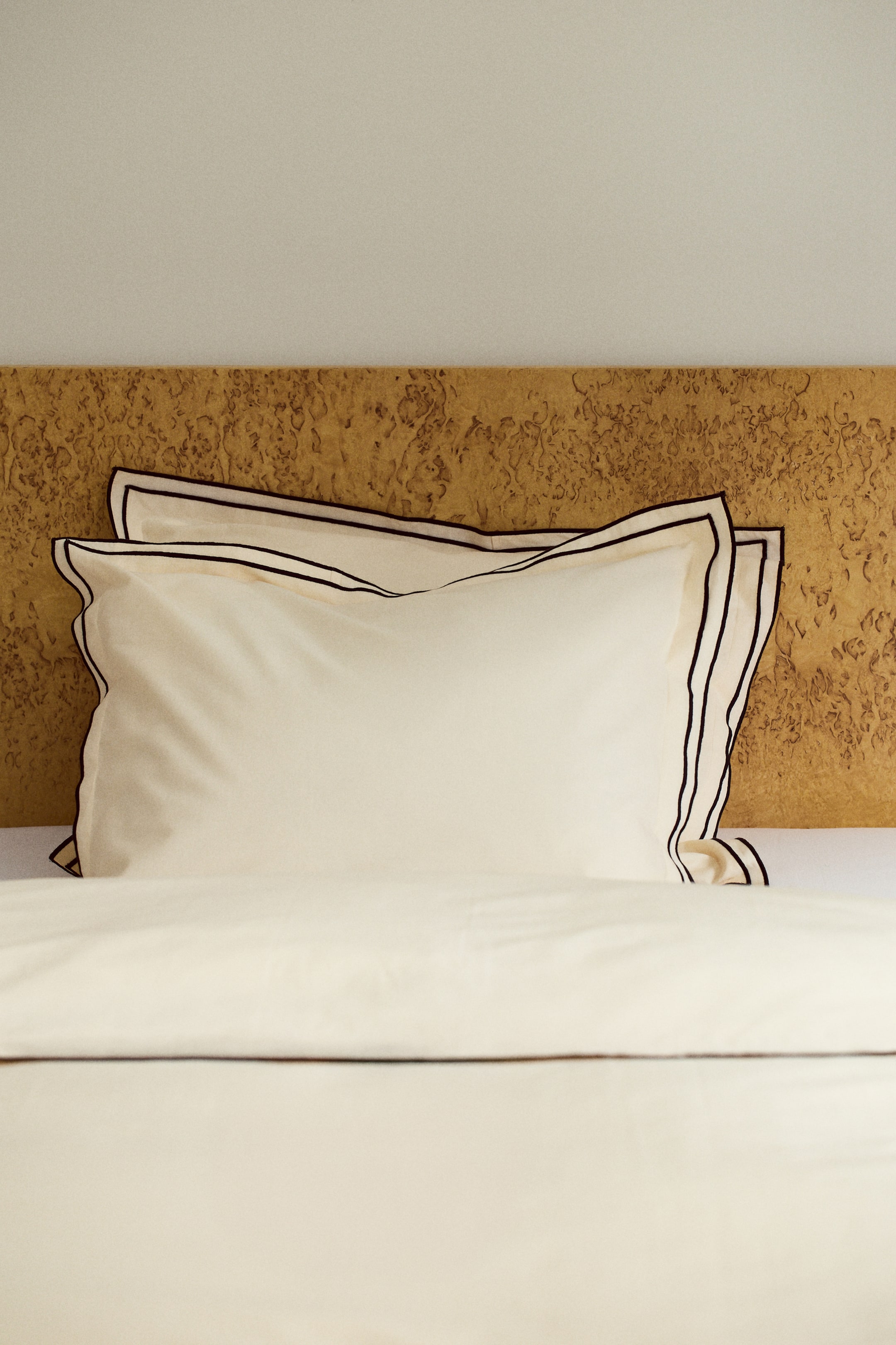 Grösseres Bild ansehen: Cremefarbene Bettwäsche zeigt zwei Kissen, die jeweils mit schwarzen Paspeln entlang ihrer geflanschten Kanten versehen sind und leicht nach links geneigt sind. Ein passendes cremefarbenes Flachbettlaken oder eine Bettdecke, ebenfalls mit schwarzen Paspeln entlang der oberen Kante verziert, liegt flach unter den Kissen.