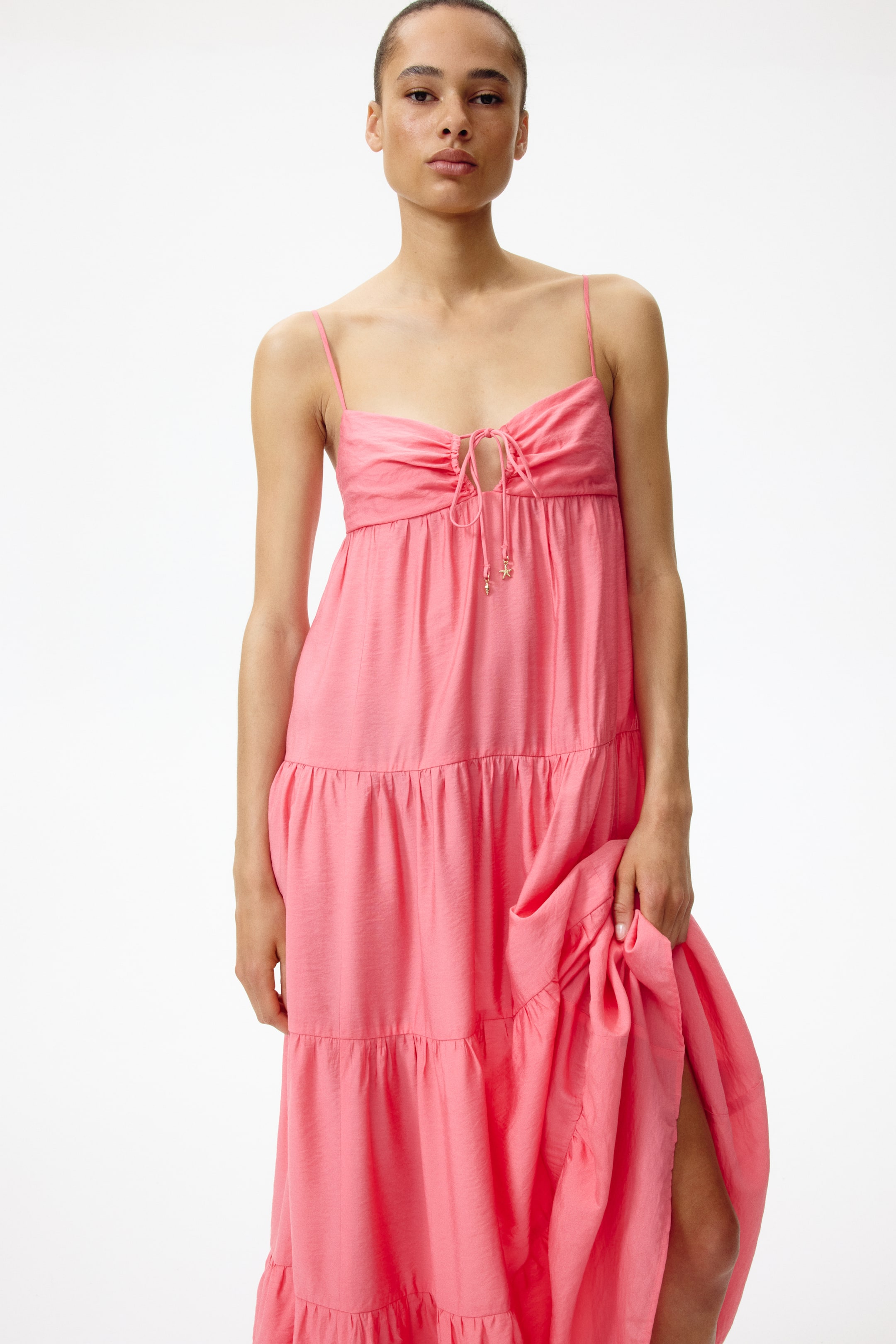 Ver imagen más grande: Un vestido maxi de color coral rosa con capas, que presenta finos tirantes de espagueti y un busto fruncido, asegurado con un cierre de cordón adornado con pequeños dijes. La larga y fluida falda muestra múltiples capas, y una parte está levantada, revelando una pierna.