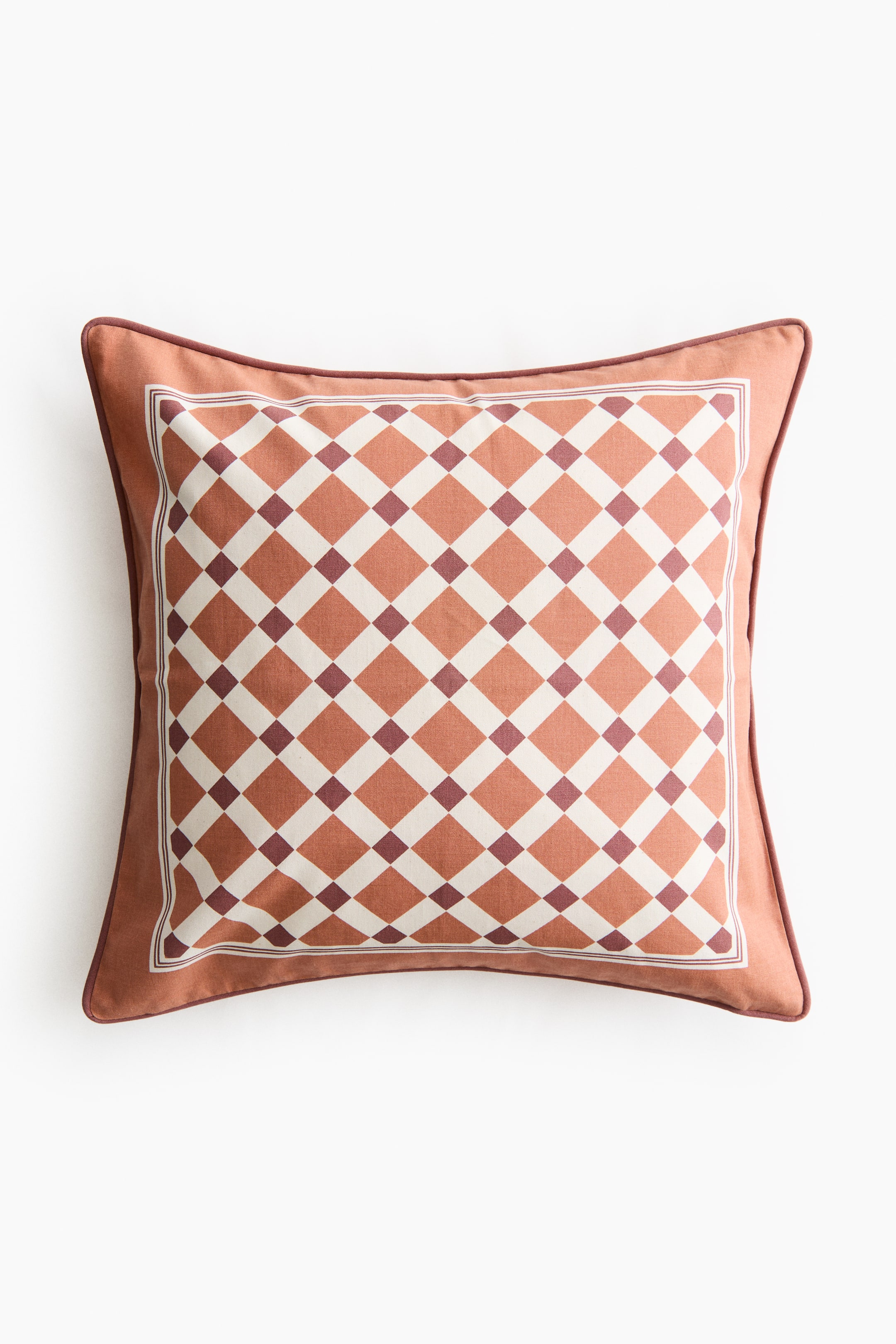 Voir une image plus grande: Un coussin carré présente sa face avant, revêtu d’un tissu orange atténué avec un passepoil brun rougeâtre foncé le long de ses bords. Le centre du coussin affiche un motif en quadrillage losangé en blanc cassé, orange atténué et brun rougeâtre foncé, encadré par une fine bordure.