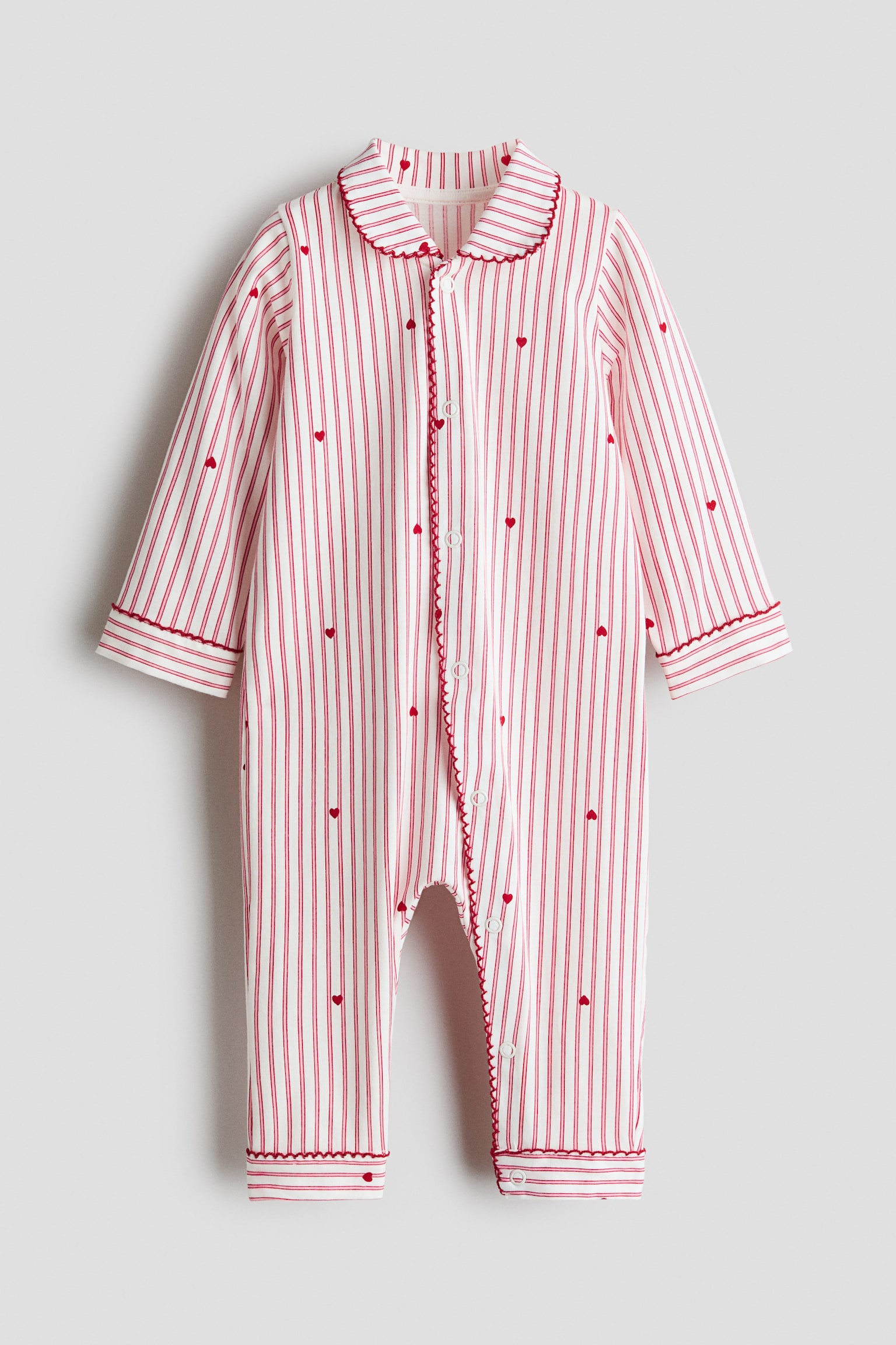 A long-sleeved infant sleepsuit is shown from the front, featuring white cotton jersey fabric patterned with vertical red stripes and tiny red hearts. The sleepsuit has a rounded collar, a snap-button placket running down the front and continuing along one leg, and red picot trim detailing the collar, placket, cuffs, and leg openings.