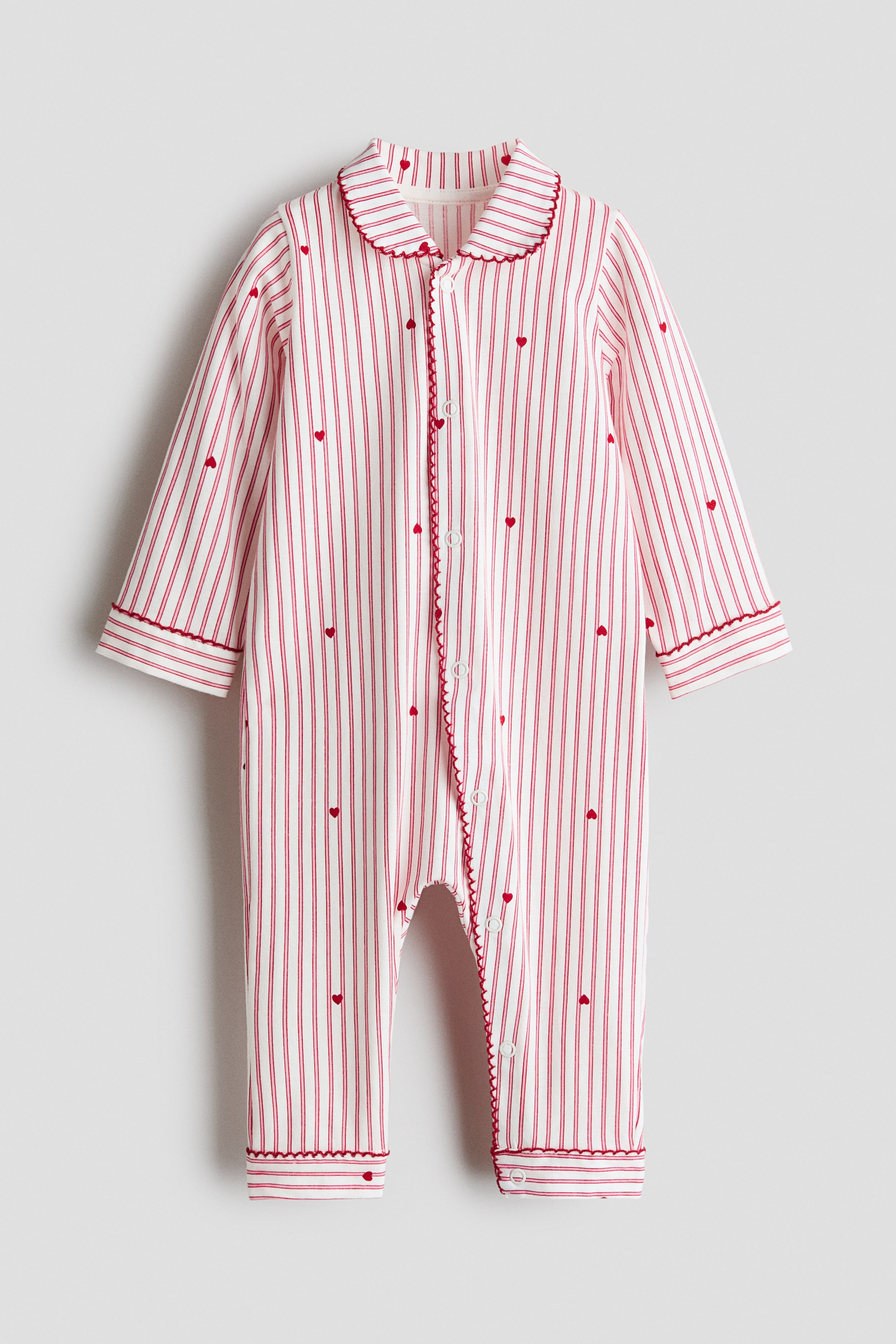 View larger image: A long-sleeved infant sleepsuit is shown from the front, featuring white cotton jersey fabric patterned with vertical red stripes and tiny red hearts. The sleepsuit has a rounded collar, a snap-button placket running down the front and continuing along one leg, and red picot trim detailing the collar, placket, cuffs, and leg openings.