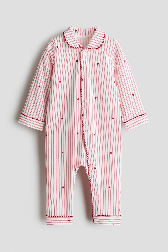 A long-sleeved infant sleepsuit is shown from the front, featuring white cotton jersey fabric patterned with vertical red stripes and tiny red hearts. The sleepsuit has a rounded collar, a snap-button placket running down the front and continuing along one leg, and red picot trim detailing the collar, placket, cuffs, and leg openings. - 1