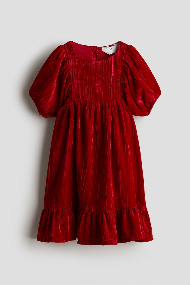 Un vestido de terciopelo rojo aplastado se exhibe desde el FRENTE, con un escote redondo y mangas cortas abullonadas con puños elásticos. El corpiño muestra pliegues verticales, y se forman frunces en la cintura y por encima del dobladillo, creando una falda de capas. Un cierre de botones es visible en el escote trasero. - 1