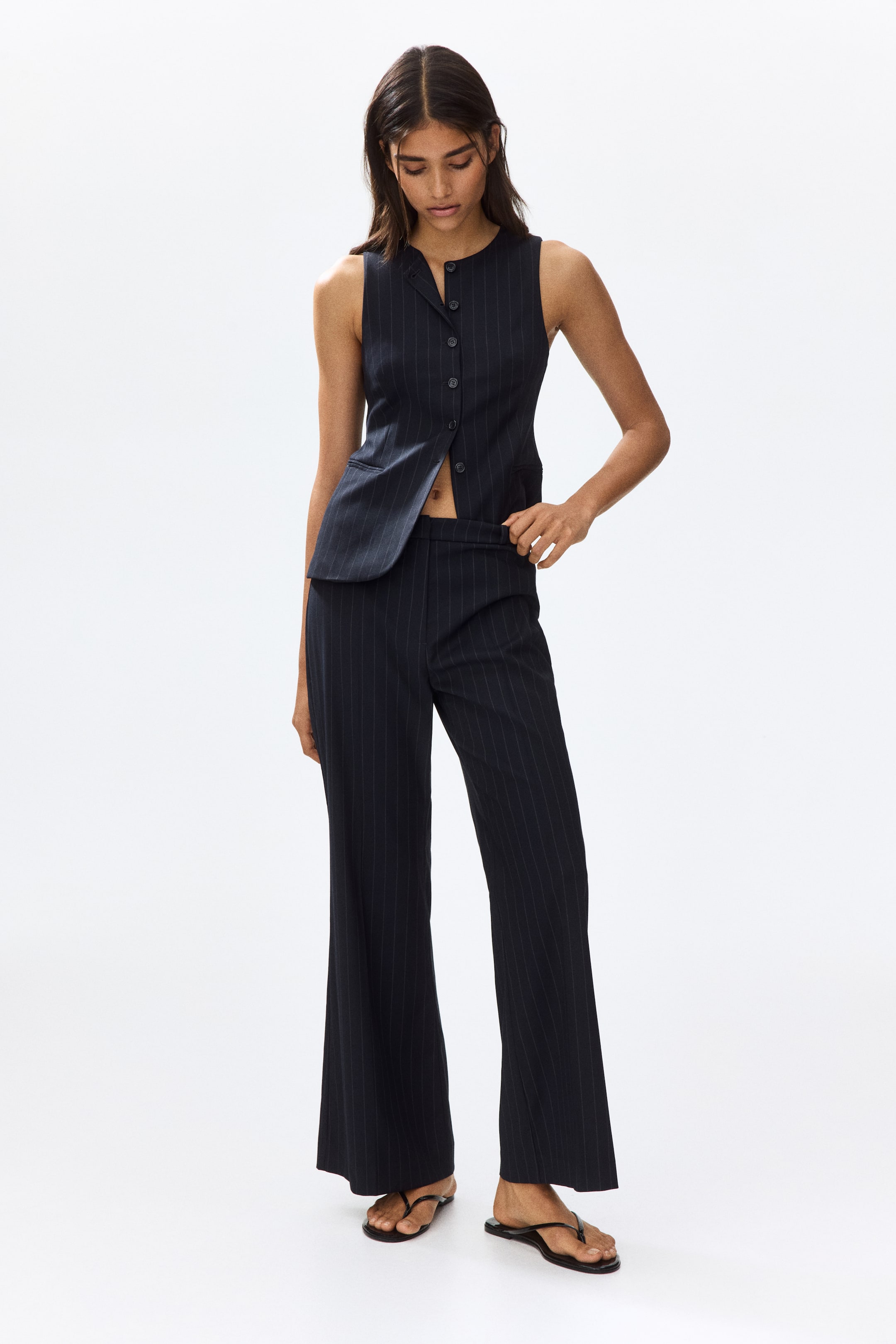 View larger image: Presenting a dark pinstriped waistcoat paired with matching trousers, showcased from the front. The sleeveless waistcoat boasts a band collar, a button-front closure, and an asymmetrical hemline. Meanwhile, the trousers feature a wide-leg silhouette.