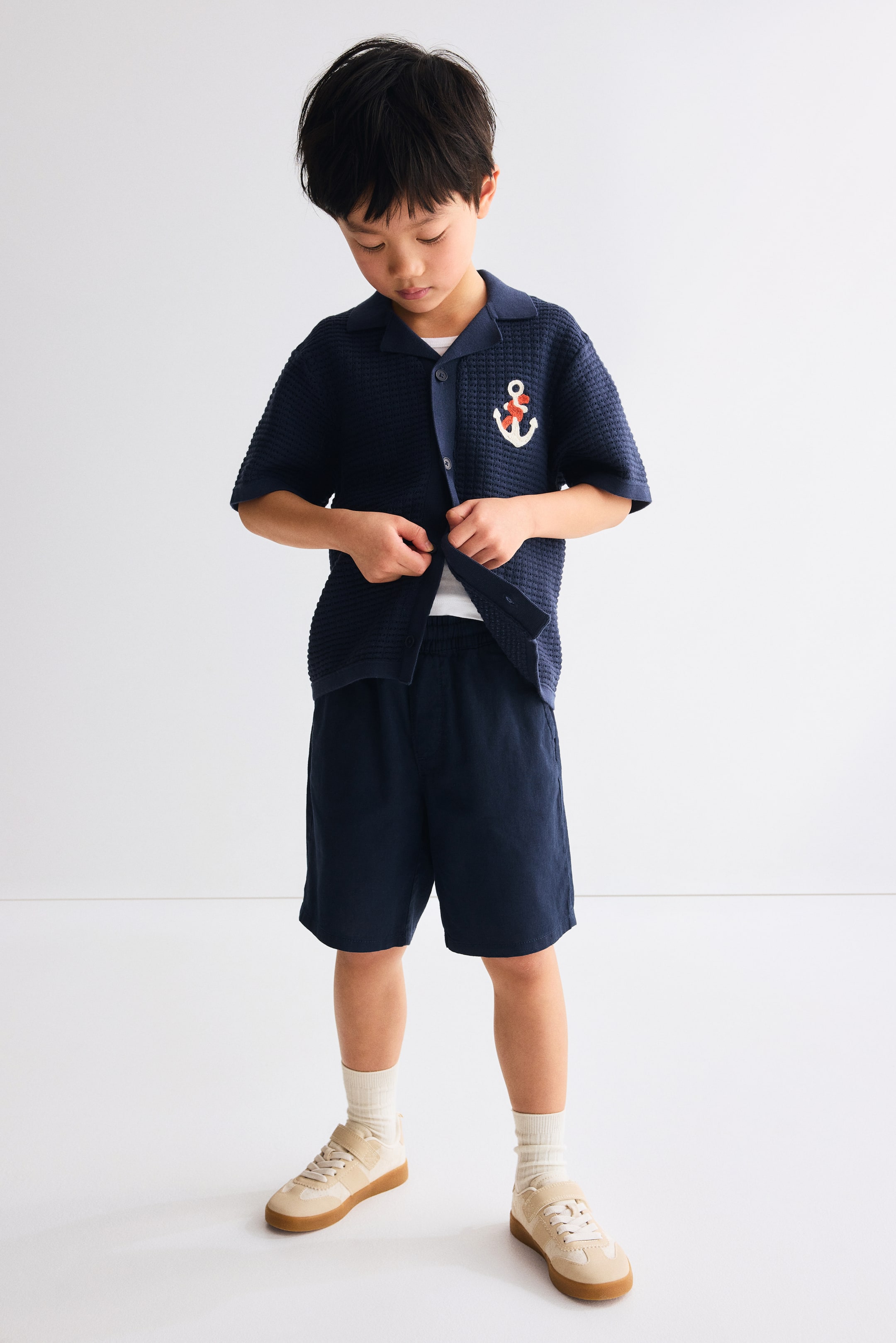 View larger image: A navy blue short-sleeved collared shirt, featuring a textured waffle knit and an embroidered red and white anchor on the left chest, is worn open over a white t-shirt. It's paired with navy blue shorts, light beige sneakers with velcro straps, and cream crew socks.