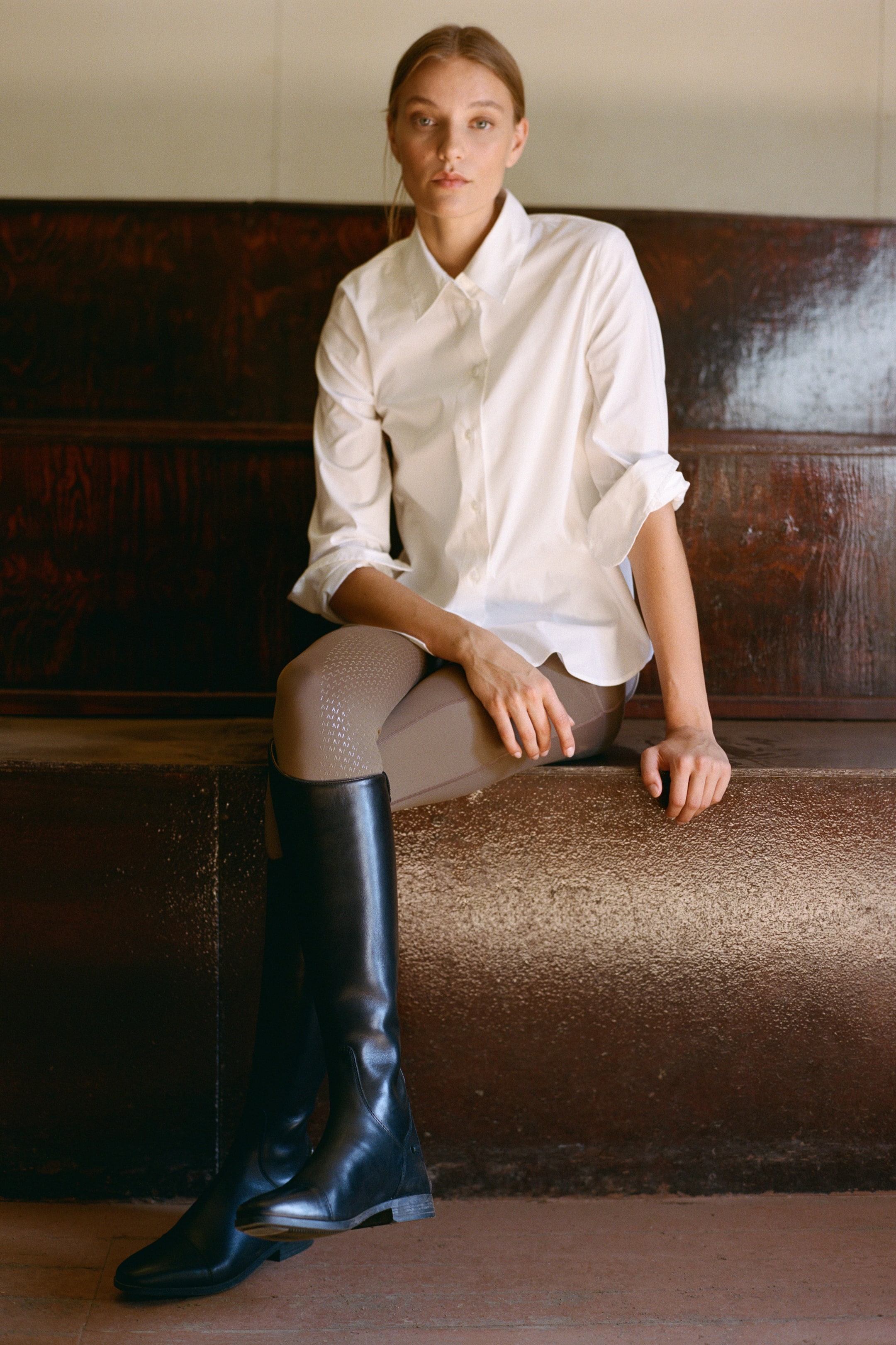 View larger image: A model is sitting facing forward, wearing taupe-coloured riding tights featuring a textured silicone grip pattern on the inner thighs. The fitted tights are styled with a white button-up shirt and tall black leather riding boots.