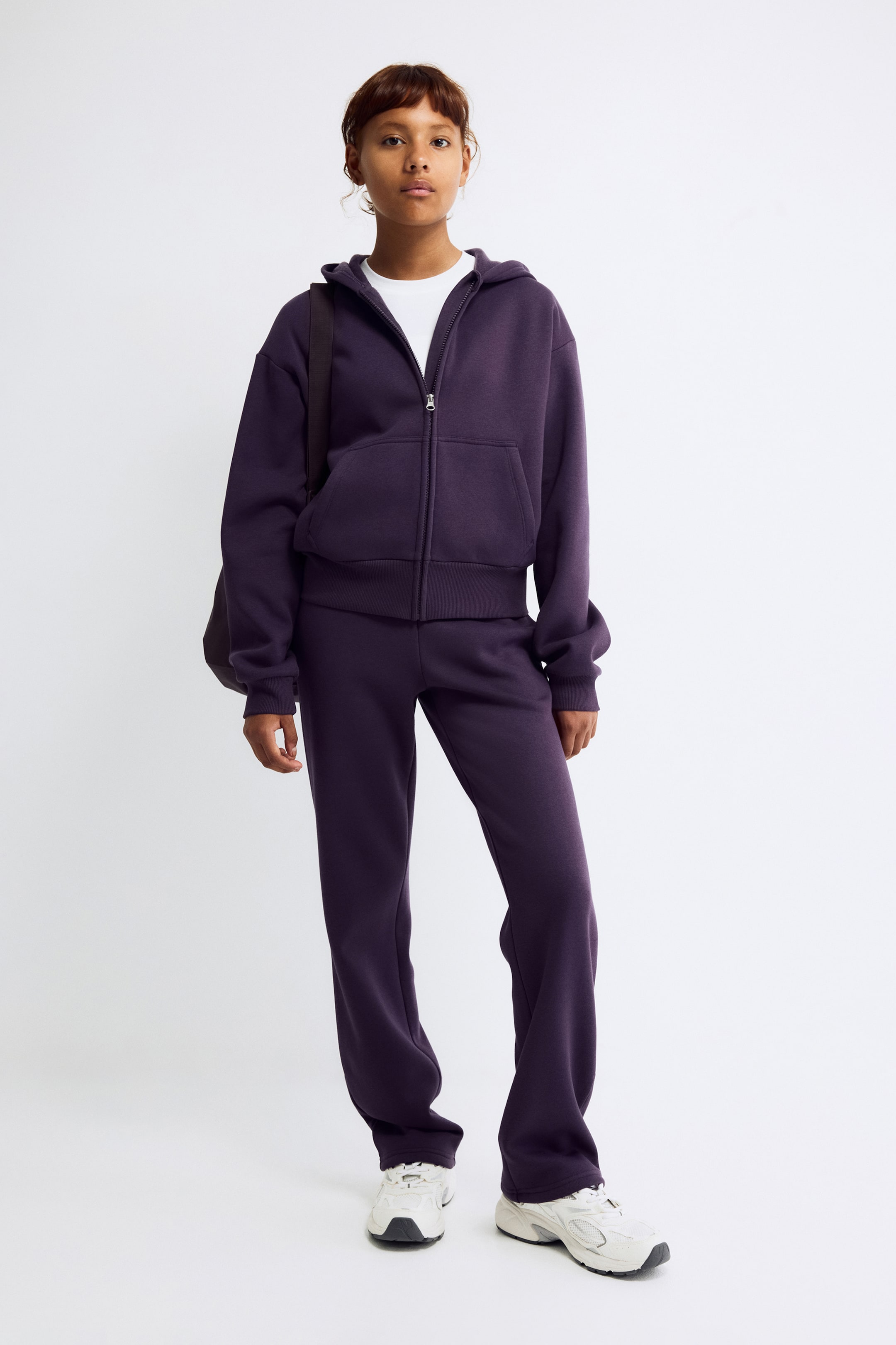 View larger image: A dark purple zip-up hoodie is shown from the front, featuring a center zipper closure, dropped shoulders, long sleeves with ribbed cuffs, and split front pockets. The hoodie is layered over a white T-shirt and paired with matching dark purple sweatpants.