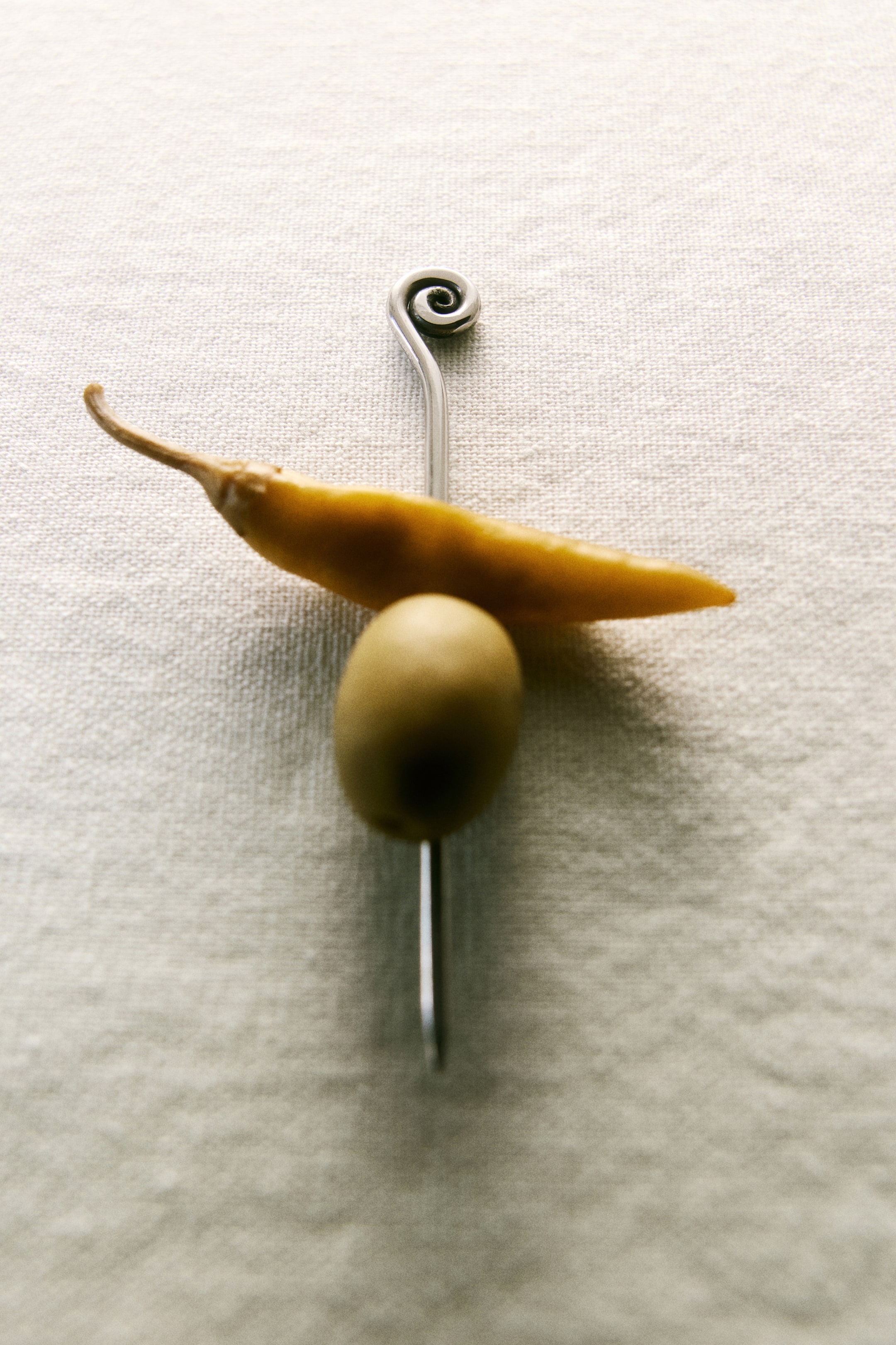 Ver imagen más grande: Un pincho de cóctel de tono plateado, que presenta un diseño en espiral en su parte superior, atraviesa una aceituna verde y un pequeño pimiento amarillo, mostrado desde una perspectiva cenital.