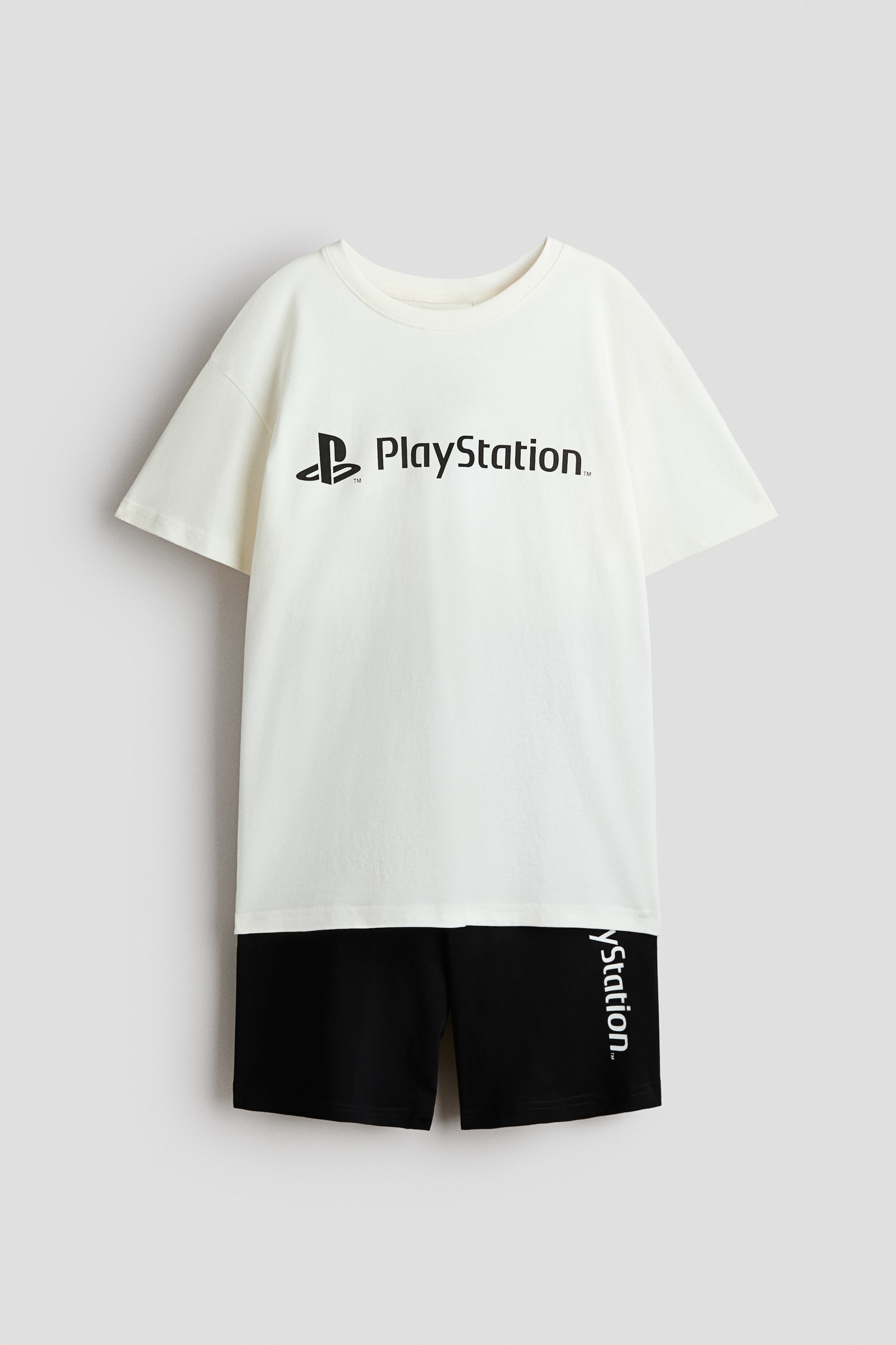 Büyük resmi görüntüle: Önden gösterilen pijama takımı, siyah PlayStation logosuna sahip krem rengi bir tişört, yuvarlak nervürlü bir yaka ve düşük omuzlarla öne çıkıyor. Eşlik eden siyah diz boyu şortlar, sol bacakta dikey 'yStation' baskısını sergiliyor ve lastikli bir bele sahip.