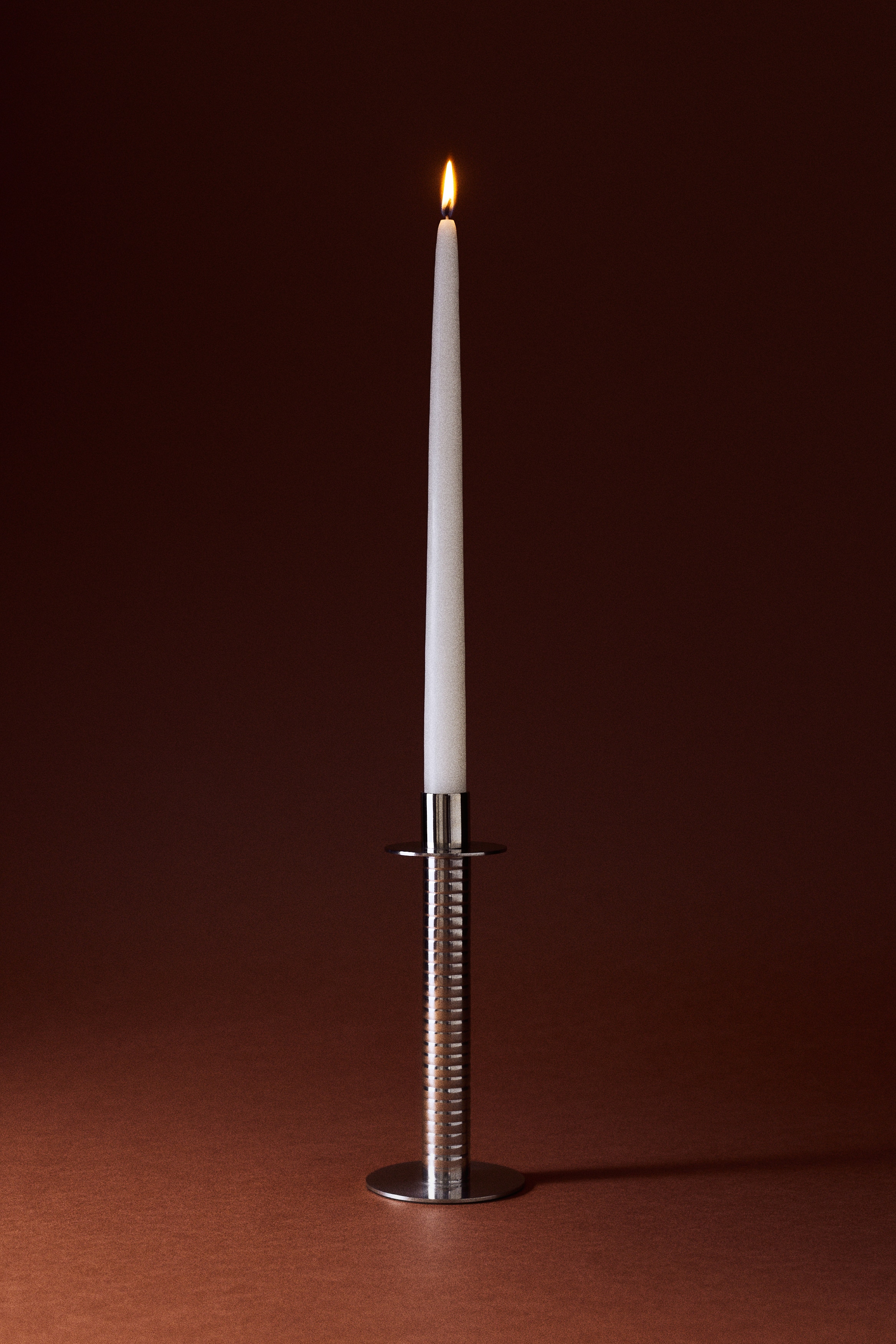Un candelabro de metal en tono plateado sostiene una vela cónica blanca. El candelabro presenta un tallo cilíndrico acanalado, una base circular plana y una bandeja circular para recoger la cera.