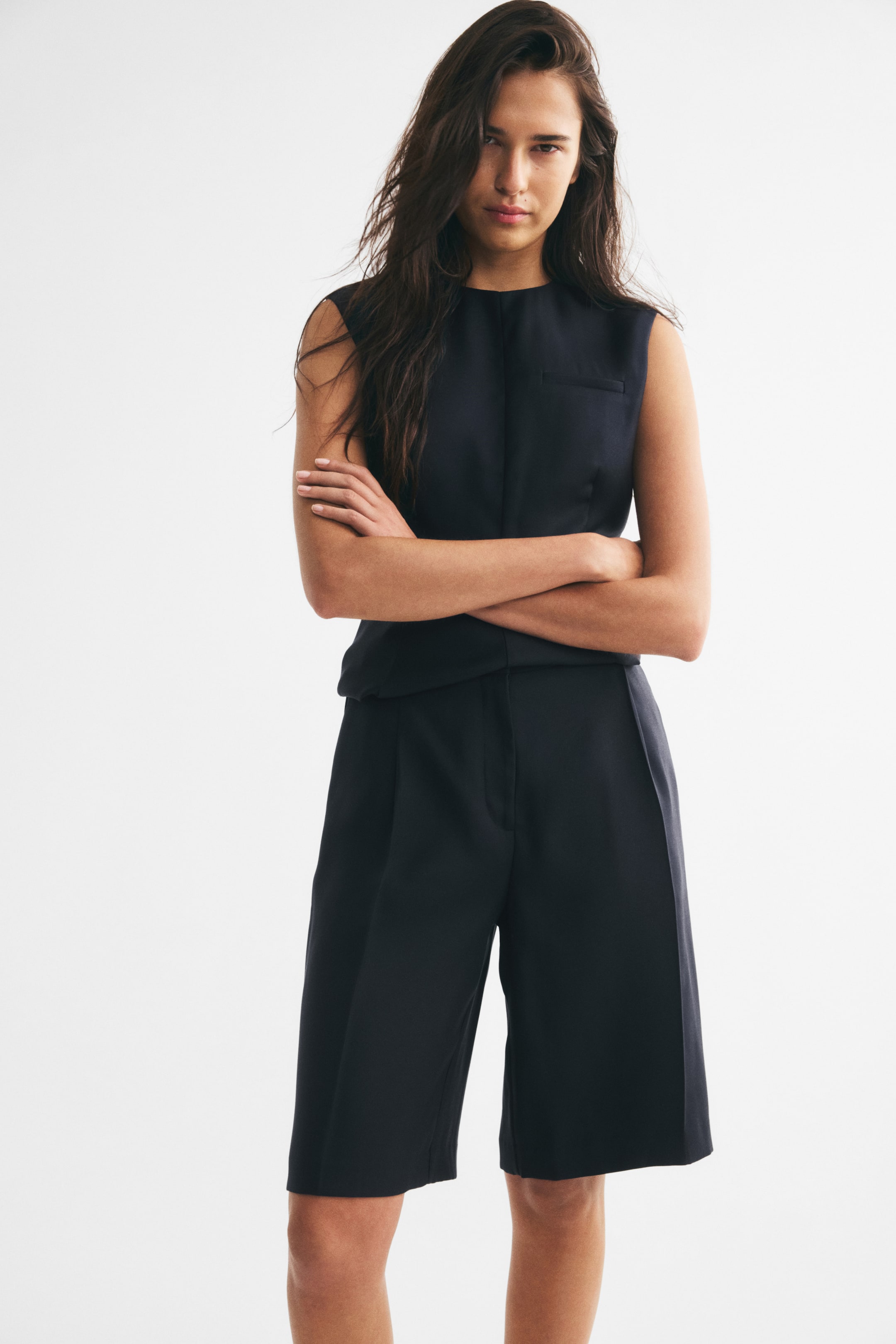 View larger image: The model is wearing knee-length tailored shorts in a dark navy basketweave, paired with a matching sleeveless top. The high-waisted shorts feature subtle front pleats and sharp center creases down the wide legs.
