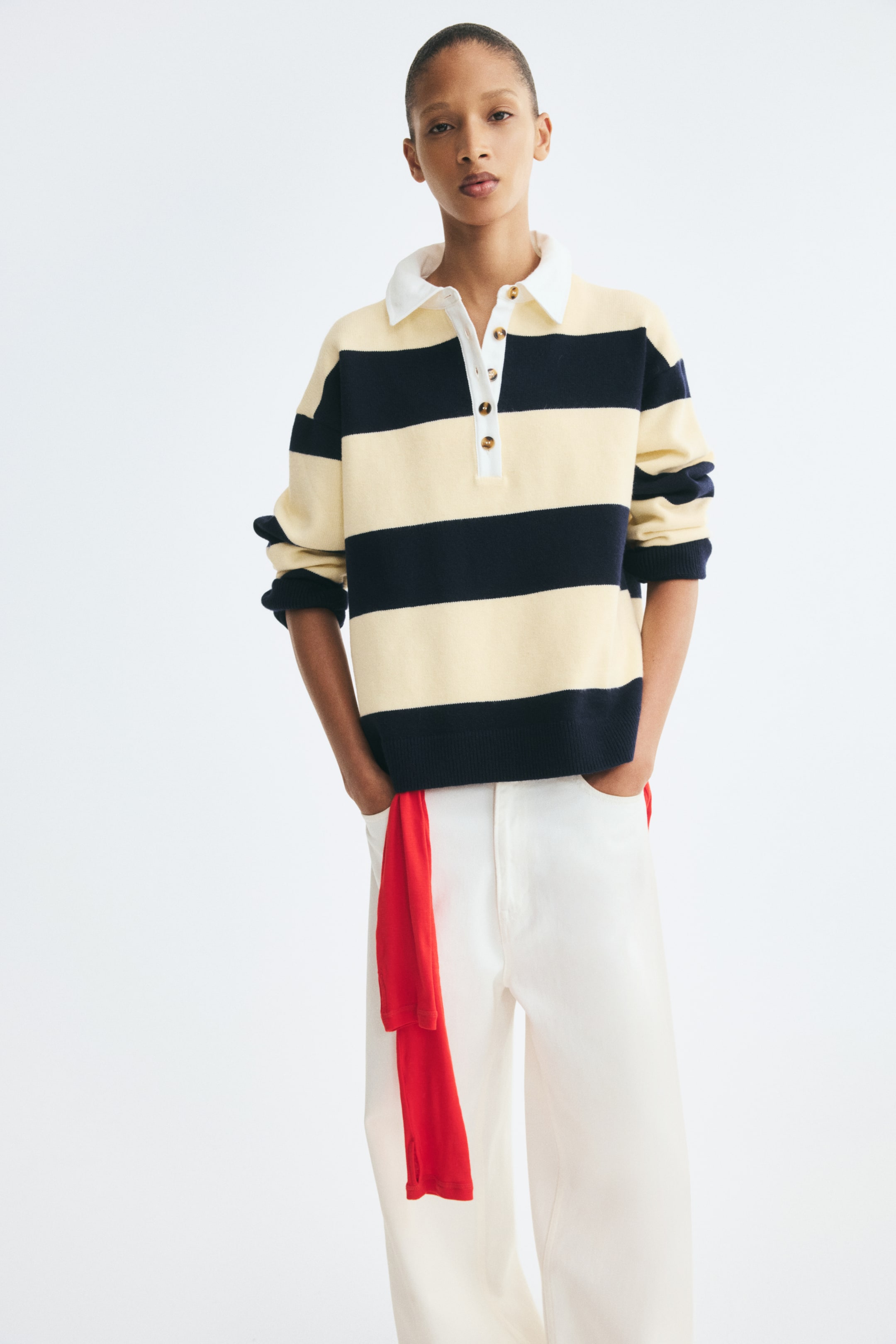 View larger image: A loose-fit knit sweater is shown from the front, featuring wide horizontal cream and navy blue stripes, a white cotton twill collar, and a button placket. The design shows dropped shoulders, long sleeves with ribbed cuffs, and a ribbed hem, and is paired with white trousers.