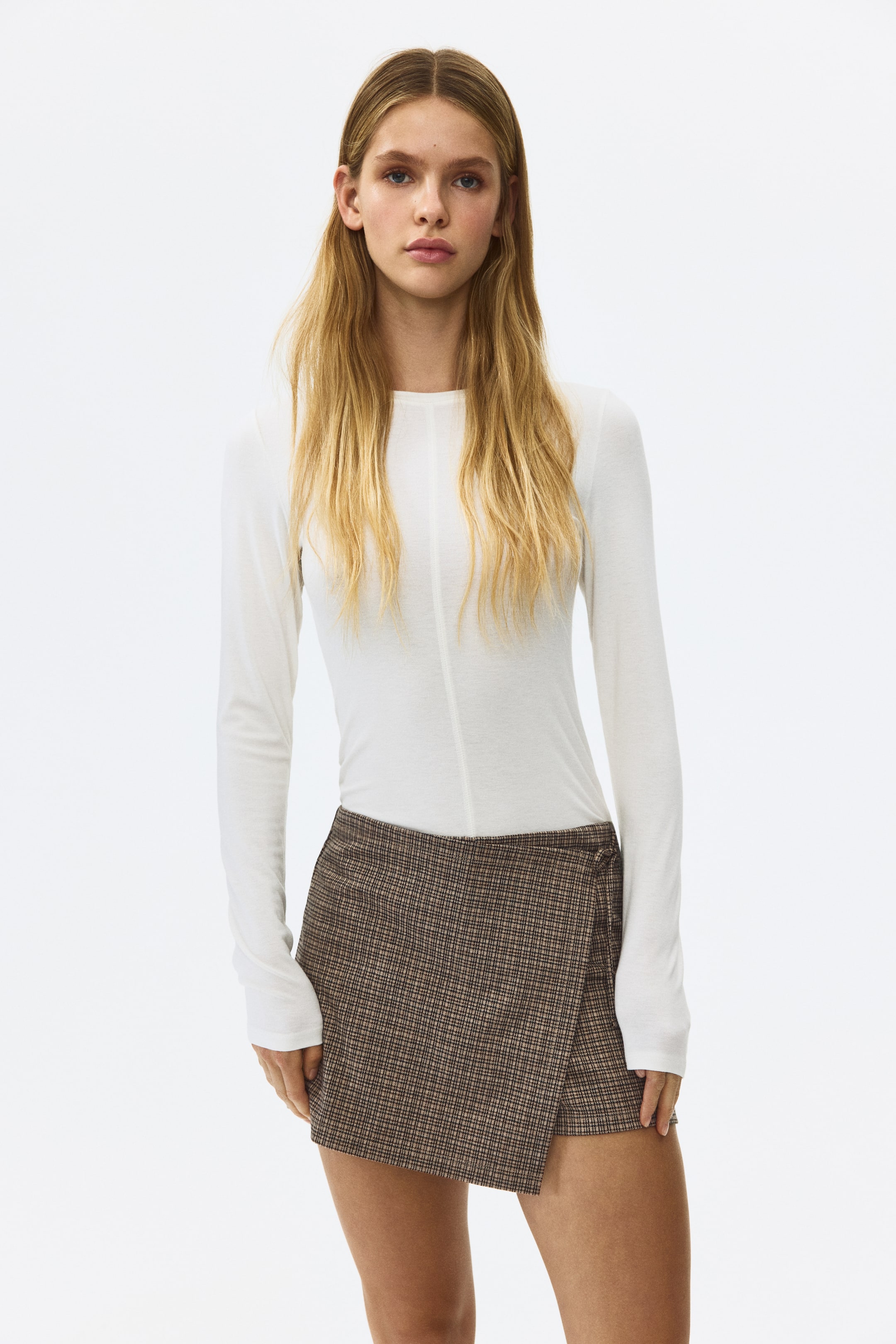 View larger image: The model is donning a plaid mini skort, characterized by a high waist and a wrapover front with slender ties on one side. The skort is paired with a white long-sleeve top.