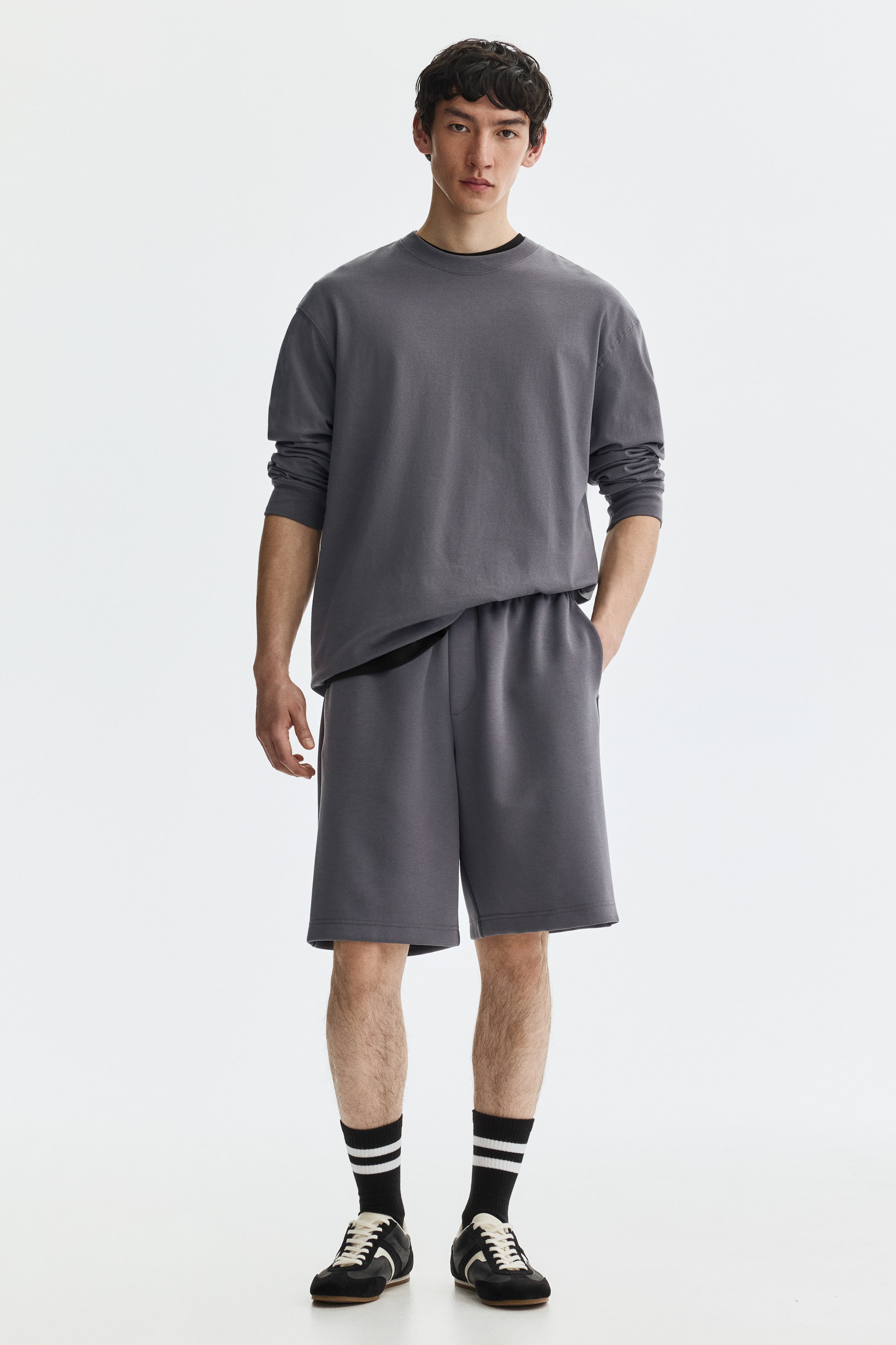 View larger image: A dark gray long-sleeve crewneck shirt is worn over a black undershirt, paired with matching dark gray shorts to complete the set. The shirt offers a relaxed fit, while the shorts feature an elastic waistband and fall just above the knees.