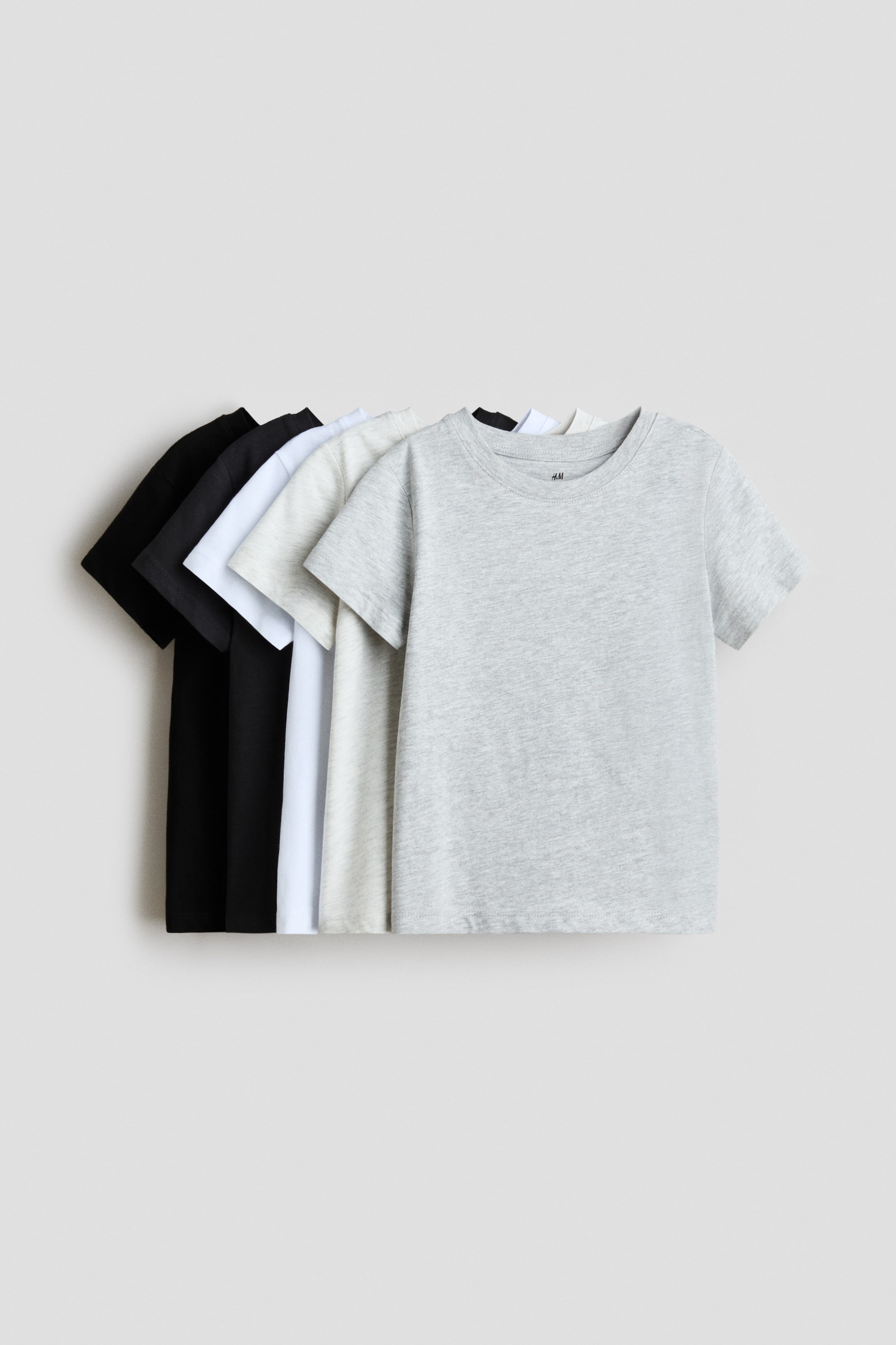 Ver imagem maior: T-shirts em jersey de algodão macio com decote redondo e bainha de corte reto são apresentadas. Cinco T-shirts surgem dispostas em sobreposição, nas cores preto, branco, cinzento claro e cinzento mesclado, com a T-shirt cinzenta mesclada virada para a frente em primeiro plano.