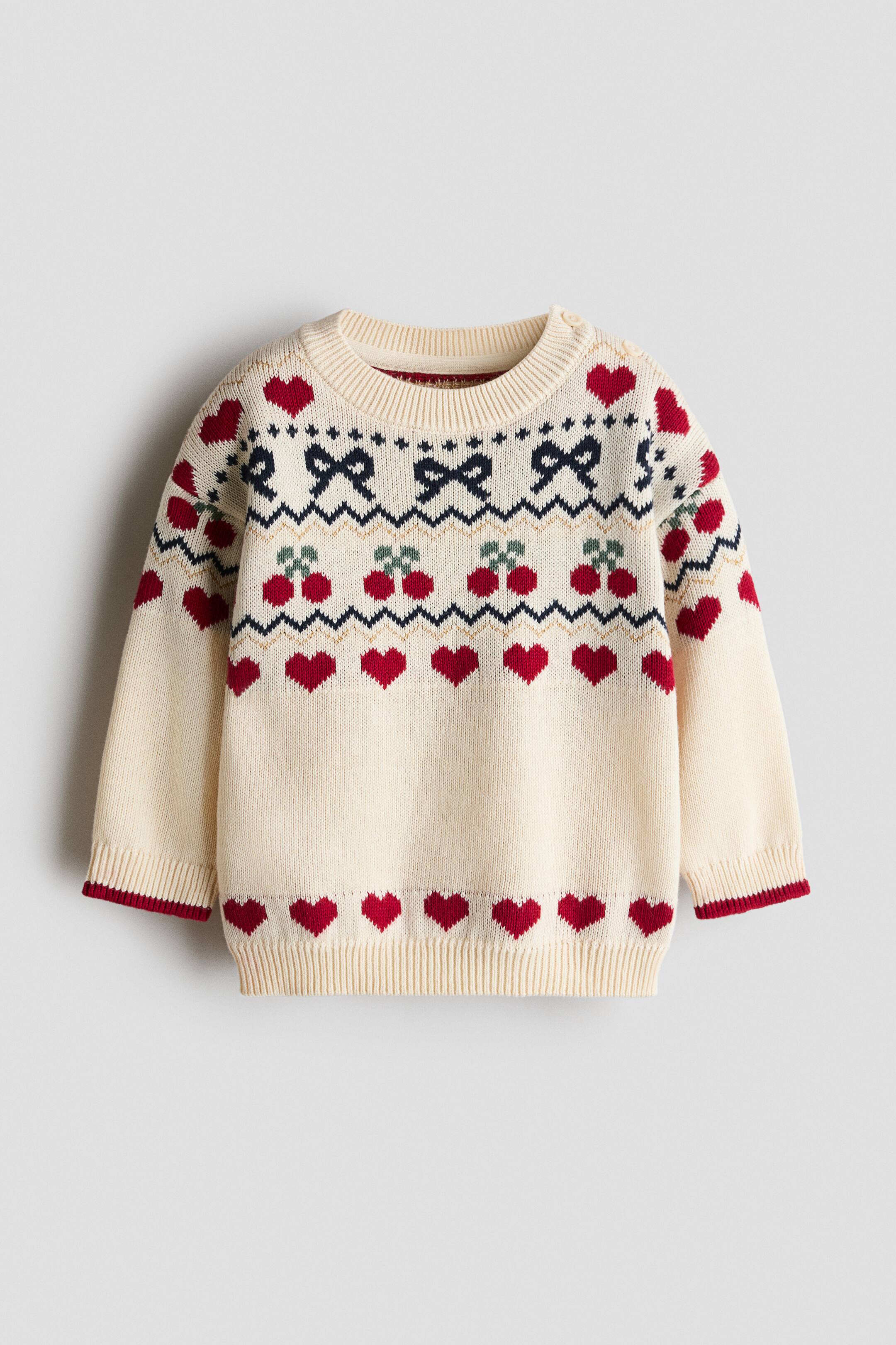 查看大圖: 一件奶油色針織毛衣正面展示,胸前及袖位飾有 Fair Isle 風格圖案,包括紅色心形、海軍藍蝴蝶結及紅色櫻桃。此款服裝採用羅紋圓領,左肩設有鈕扣開合,羅紋袖口飾以紅色滾邊,以及羅紋下擺。