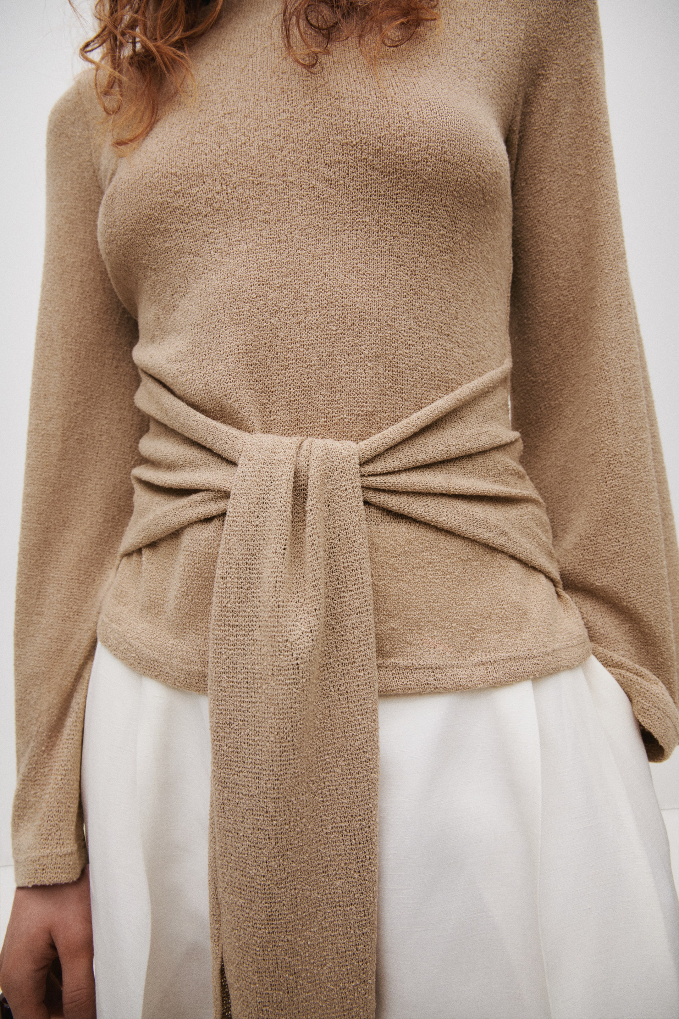 Grösseres Bild ansehen: Ein strukturiertes Strickoberteil mit einem U-Boot-Ausschnitt und langen, weiten Ärmeln wird von dem Model getragen. Lange, breite Bänder, die von den Seiten des Oberteils ausgehen, werden vorne umwickelt und gebunden.