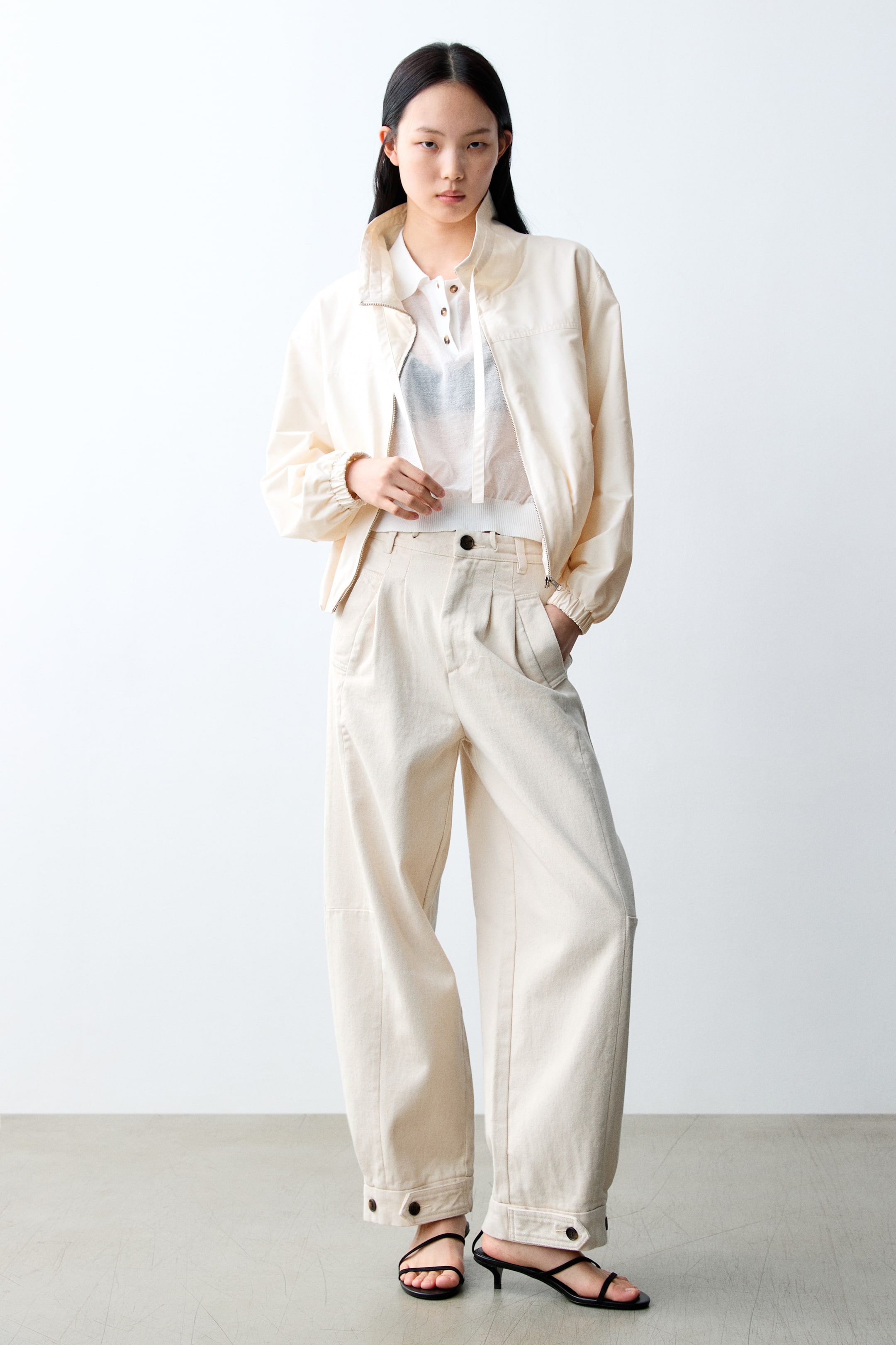 이미지 확대 보기: 크림색의 봄버 재킷이 스탠드 칼라와 주름진 커프스로 특징을 이루며, 리브드 헴이 있는 시어 화이트 카라 폴로 셔츠 위에 레이어드되어 있습니다. 크림색 와이드 레그 바지는 주름과 버튼식 발목 커프스로 완성되며, 블랙 스트래피 키튼 힐 샌들과 함께 착용하여 룩을 완성합니다.