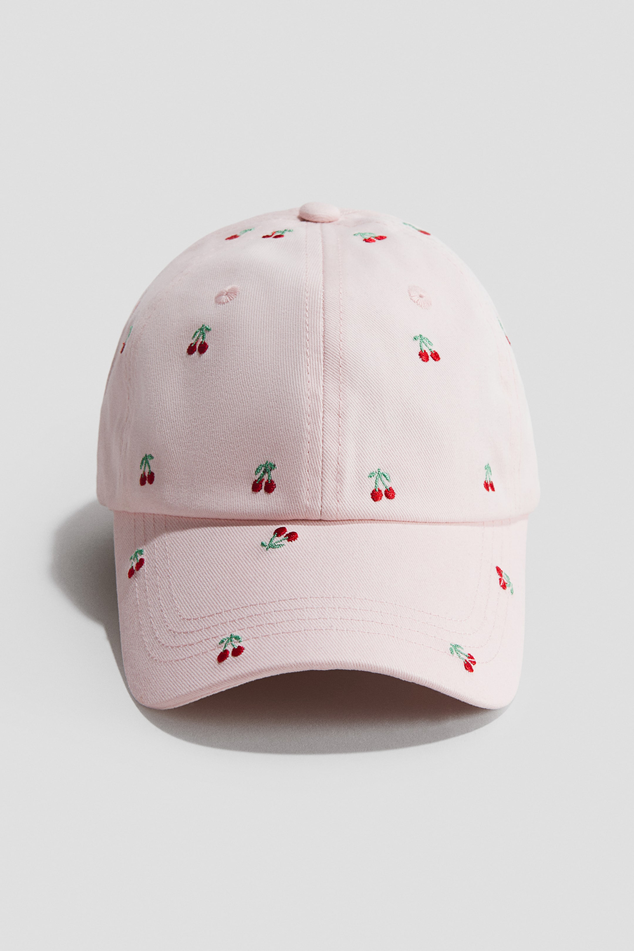 View larger image: A light pink cap is showcased FRONT, adorned with small embroidered red cherries scattered across its surface and a curved brim. It features a six-panel design, topped with a fabric-covered button.