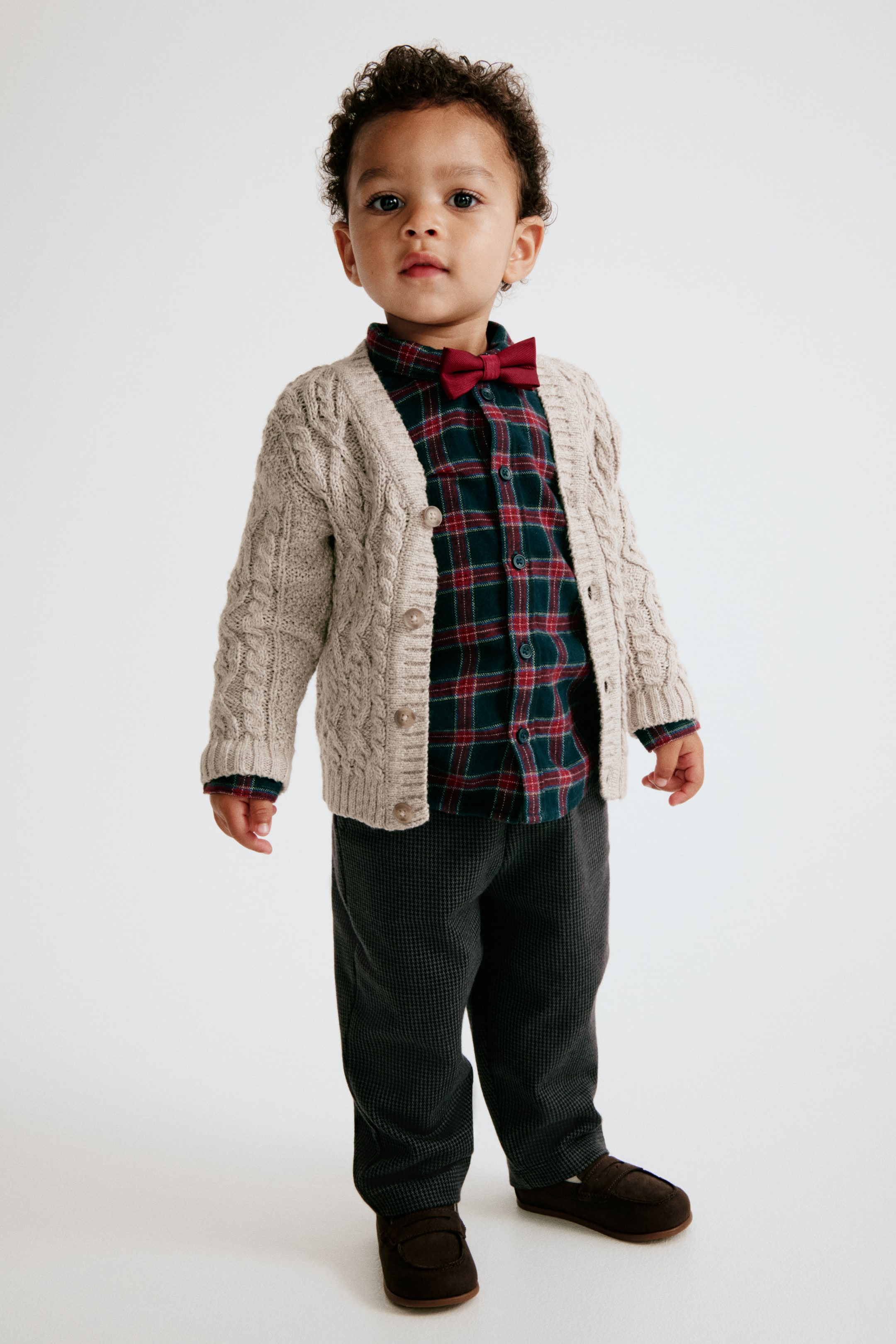 View larger image: These dark grey trousers in textured jacquard jersey are featured from the front on a toddler, showing a relaxed, straight-leg fit. The trousers are styled with a green and red plaid shirt, a beige cable-knit cardigan, and dark brown loafers.