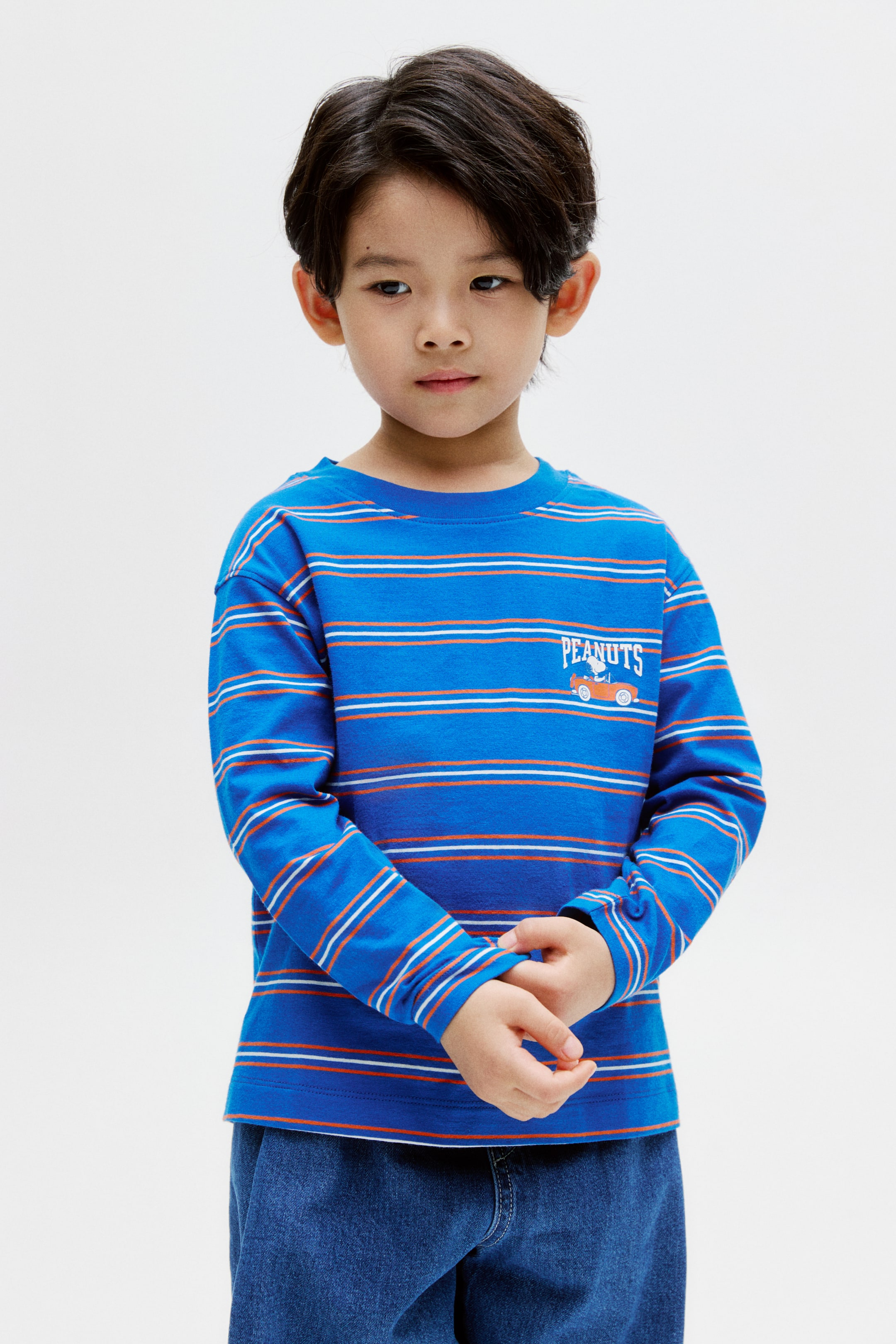拡大画像を表示: 青色の長袖クルーネックTシャツは、オレンジと白の横ストライプが特徴で、左胸には「PEANUTS」の文字と赤い車を運転するスヌーピーの小さなグラフィックが描かれています。