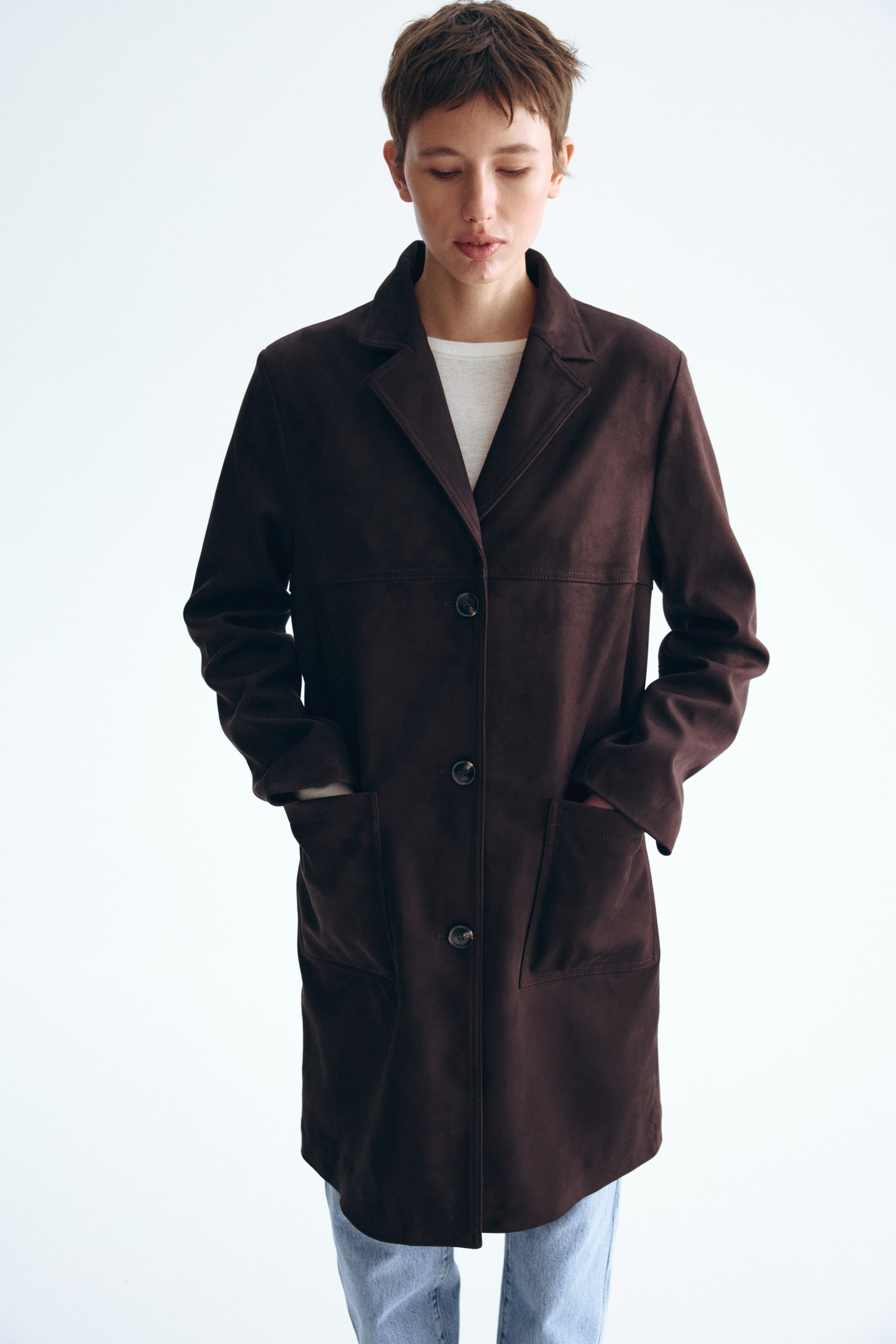 View larger image: A dark brown suede-like coat is facing front, featuring notch lapels and a three-button single-breasted fastening. It is designed with two large patch pockets and is layered over a light-coloured top.