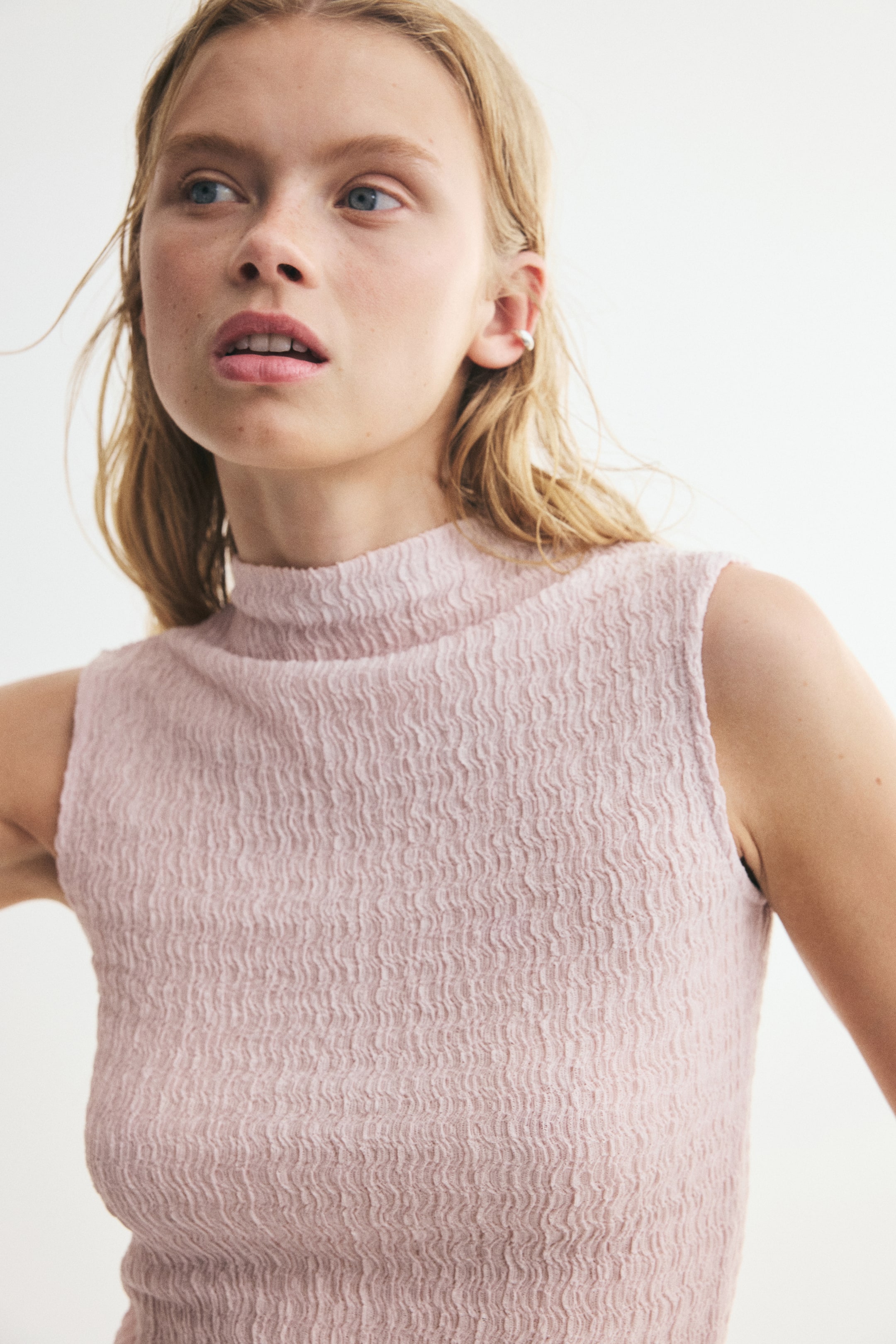 View larger image: The model is wearing a light pink sleeveless top in a crinkled jersey fabric. The top features a high neck and a slim fit, highlighting its richly textured pattern.