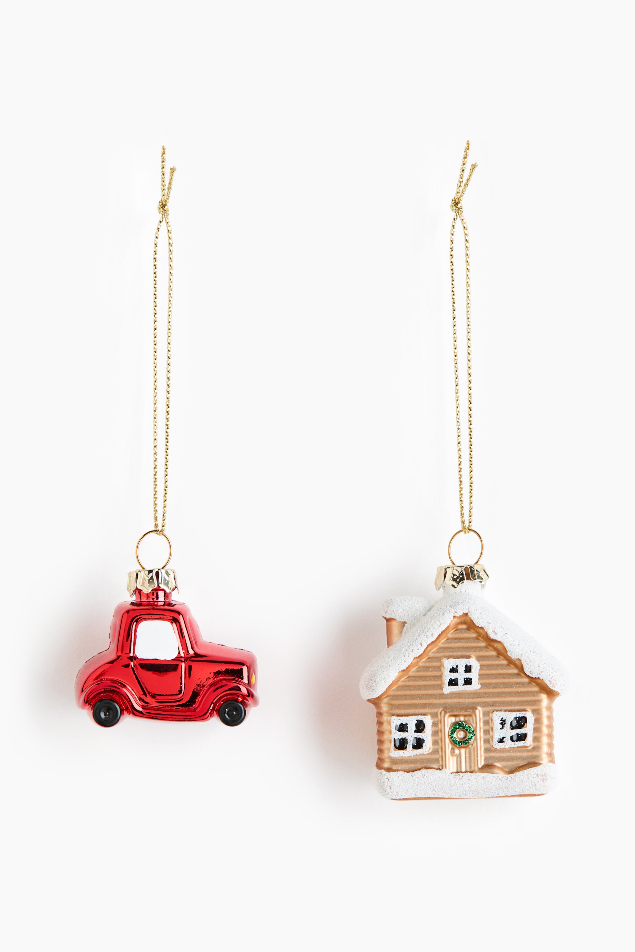 Ampliar la imagen: Dos adornos decorativos cuelgan de hilos dorados, orientados hacia el frente. El adorno de la izquierda es un auto rojo brillante, con ruedas negras y ventanas blancas, mientras que el adorno de la derecha es una casa dorada, con detalles de nieve blanca en el techo y la base, ventanas negras y una pequeña corona verde en la puerta.
